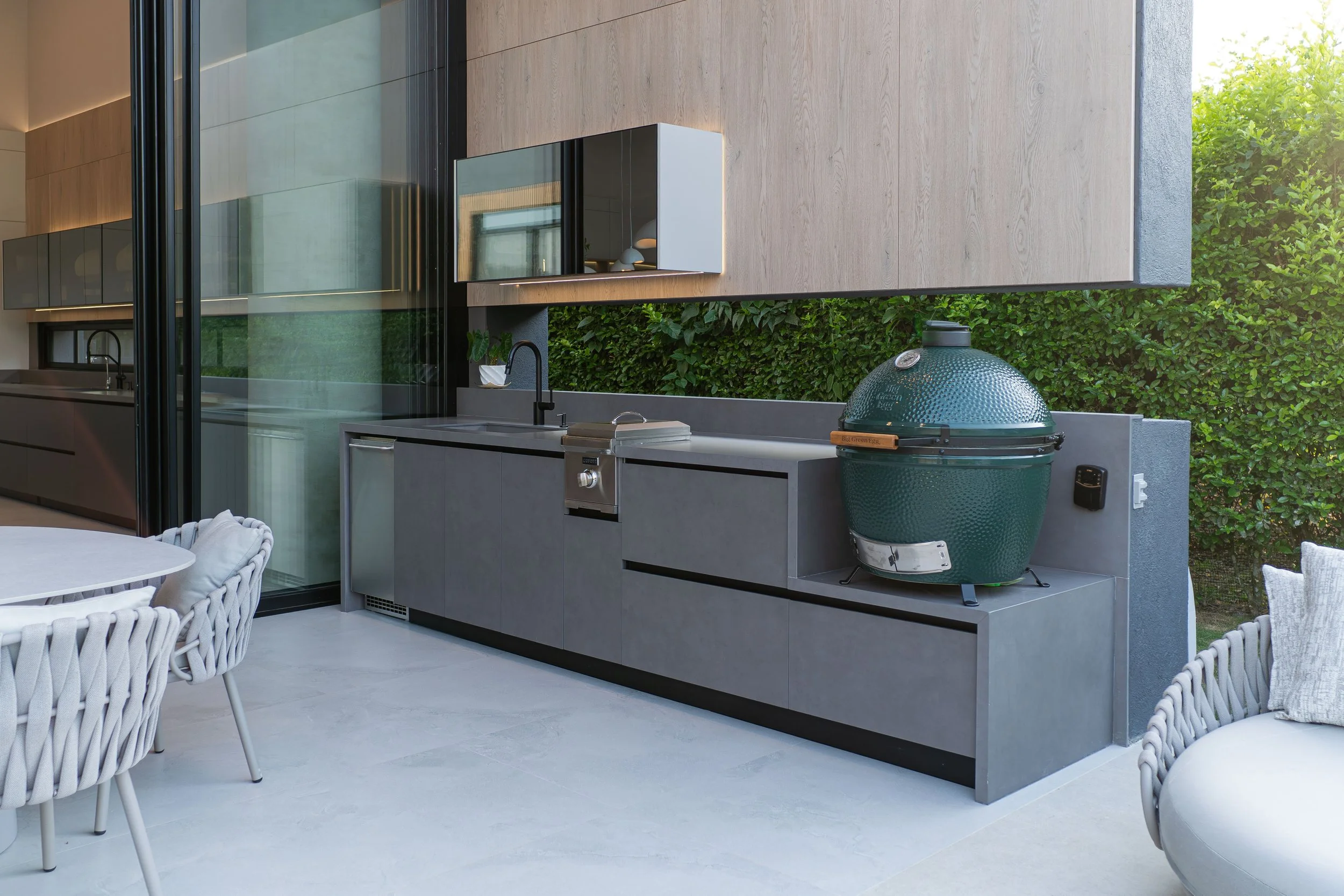 Terraza moderna con área de cocina exterior que incluye un asador verde tipo kamado, mostrador gris, fregadero y muebles de almacenamiento. Amplio espacio con sillas y cojines, rodeado de vegetación.
