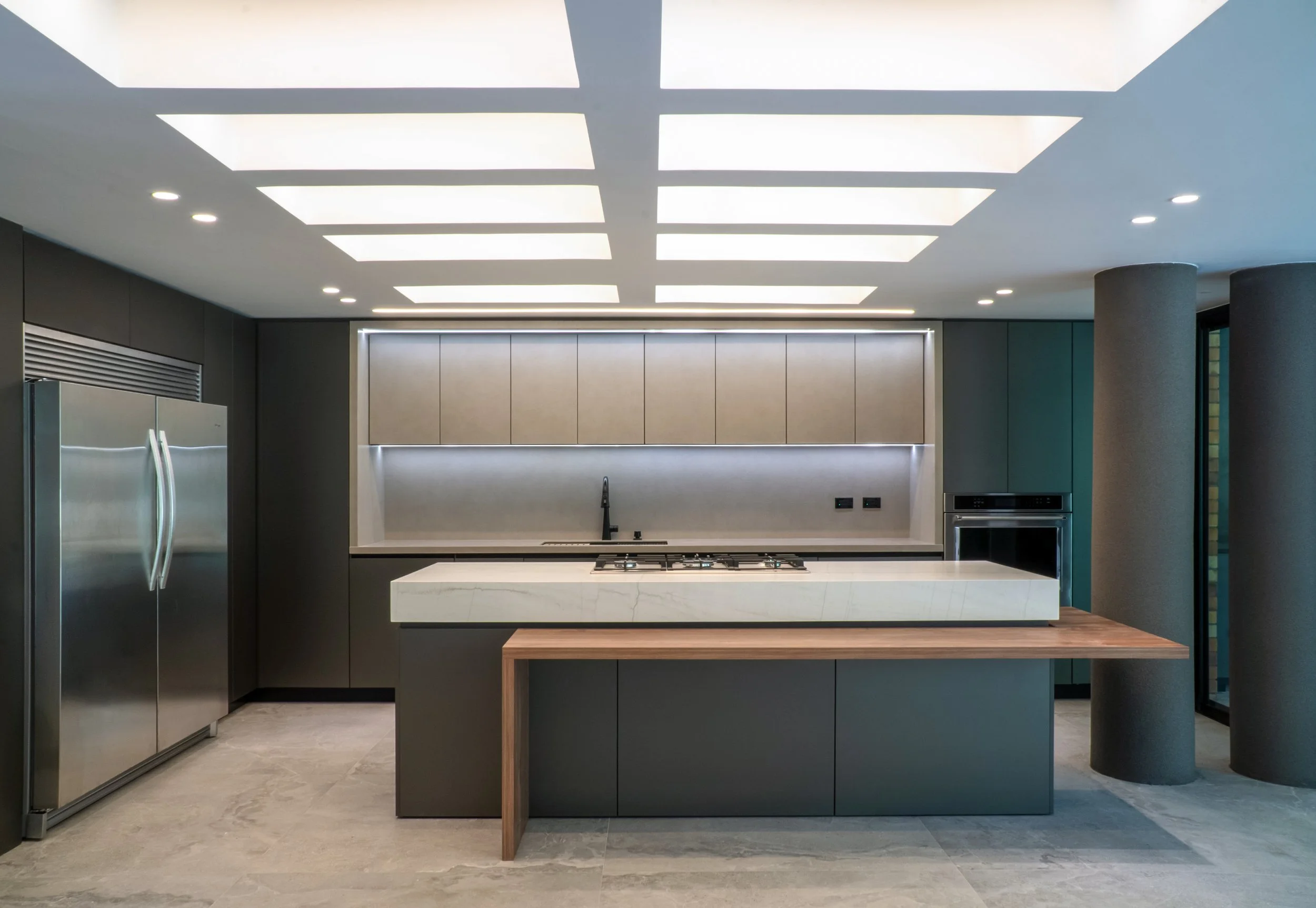 Cocina moderna con gabinetes grises, una isla con encimera de piedra, electrodomésticos de acero inoxidable y un techo con paneles de luz empotrados.