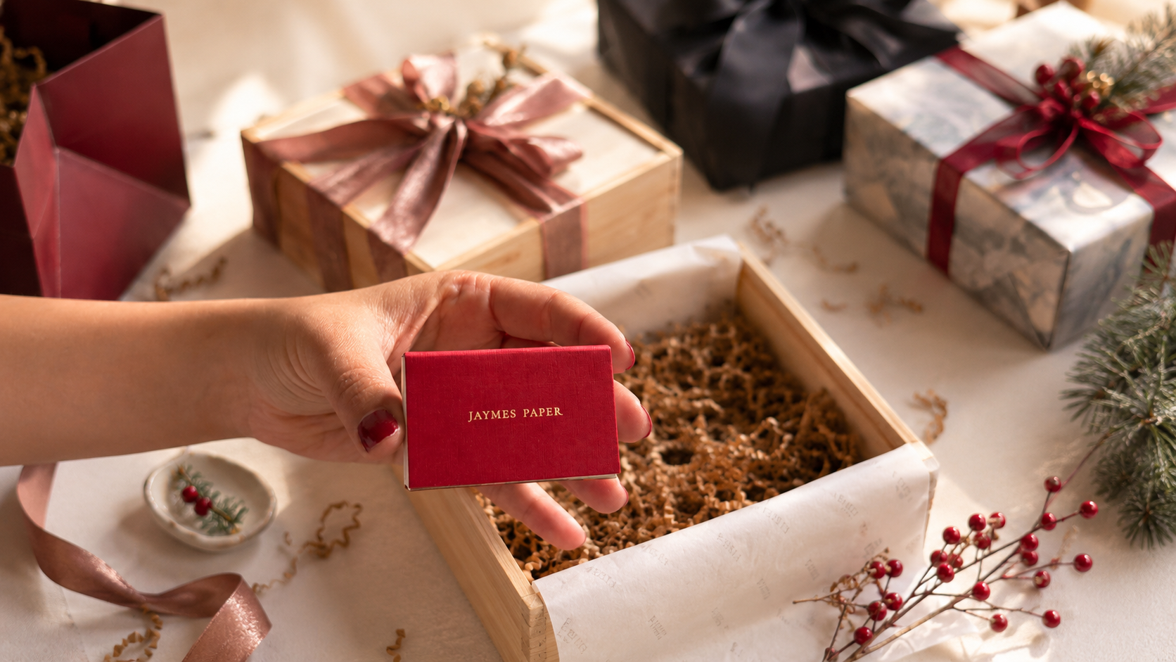 A hand placing a small gift into a wooden gift box surrounded by ribbon and holiday styling