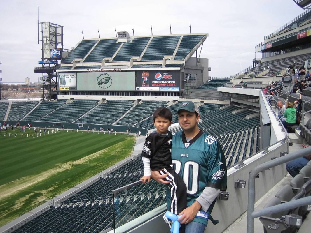 A man and a young boy in Eagles football jerseys at a football stadium, with empty seats and a field in the background.