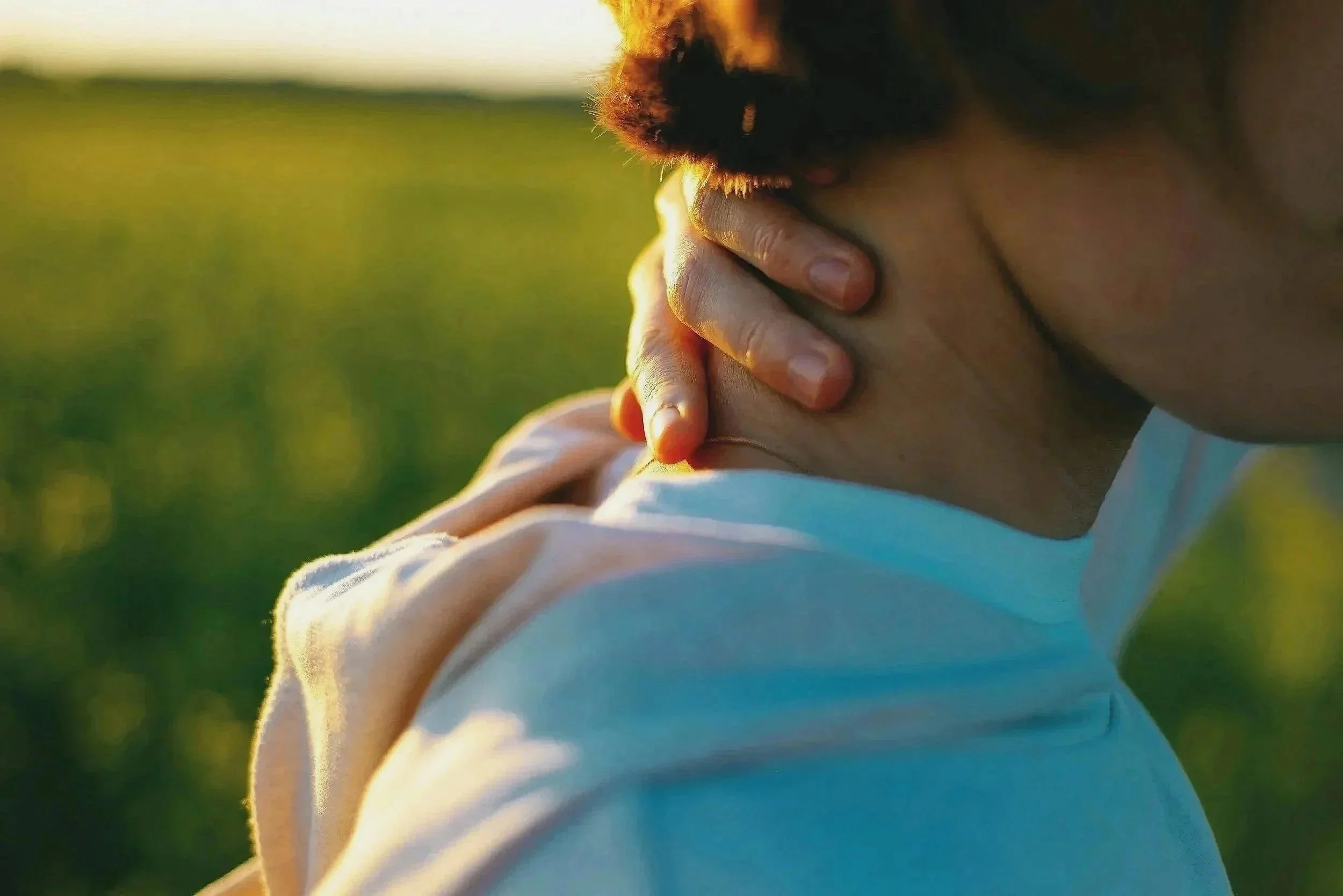 Close-up of a person with light skin, wearing a white shirt, holding their hands together near their face, with a background of green grass and trees illuminated by warm sunlight.
