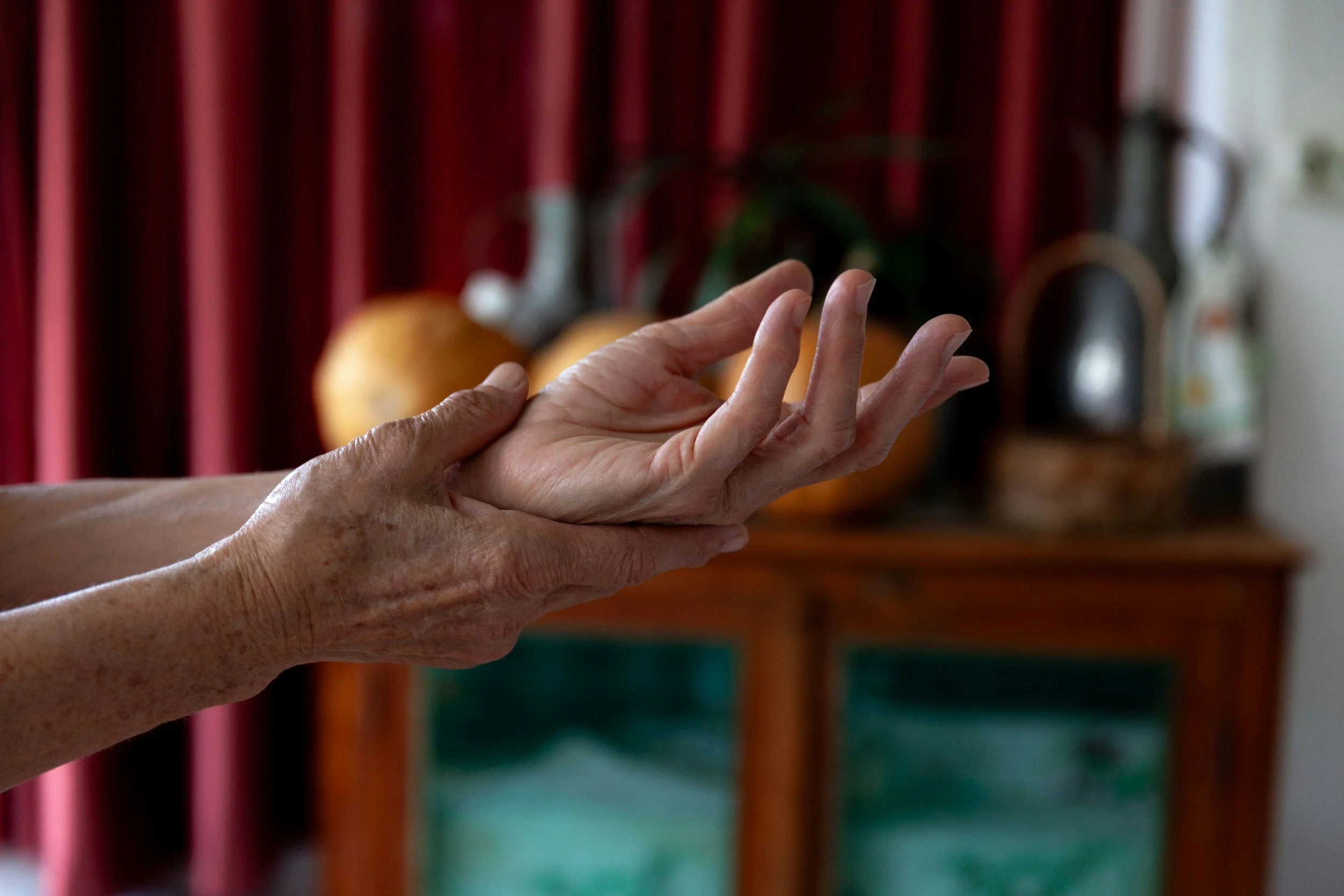 Close-up of a person's hand with fingers slightly raised, in front of a blurred background of a wooden shelf with pumpkins and various bottles. The person is showing pain in their wrist or hand.