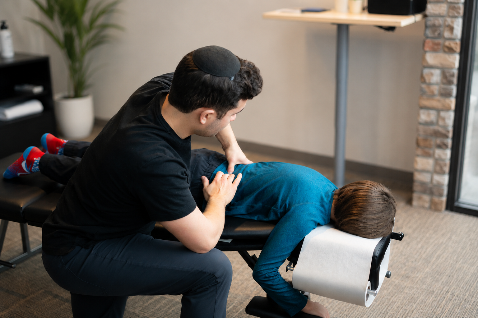 A chiropractor performing an treatment on a young boy lying face down on an examination table in a clinic or therapy room.