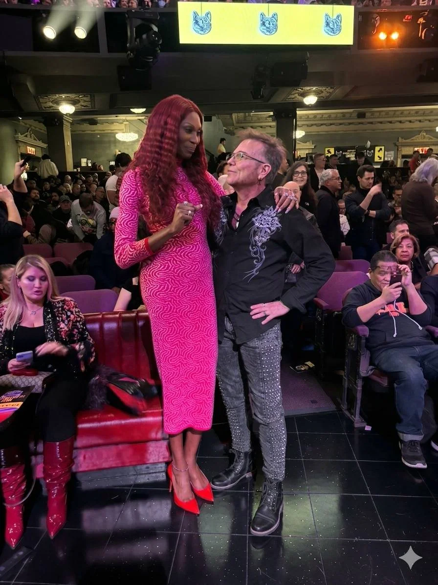 A woman with long, curly red hair wearing a pink dress and red high heels standing close to a man with glasses, wearing a black shirt with silver embroidery and patterned pants, in a crowded indoor event space. The woman has her arm around the man's shoulders, and they appear to be interacting in front of an audience.