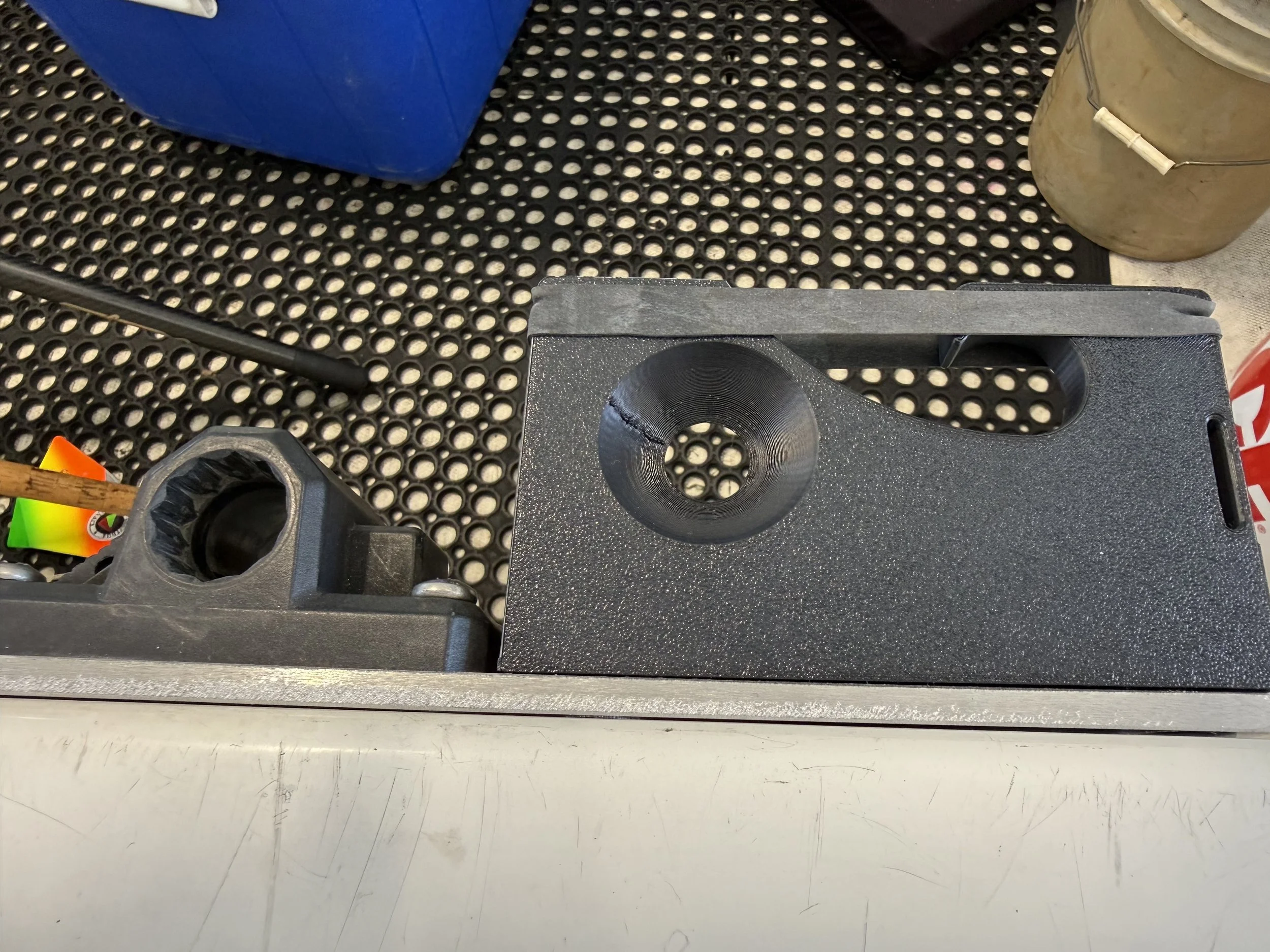 A black piece of equipment with a large circular hole on top, placed on a white surface, next to a black rubber mat with round holes, a yellow container with a handle, a blue box, and a small colorful object.