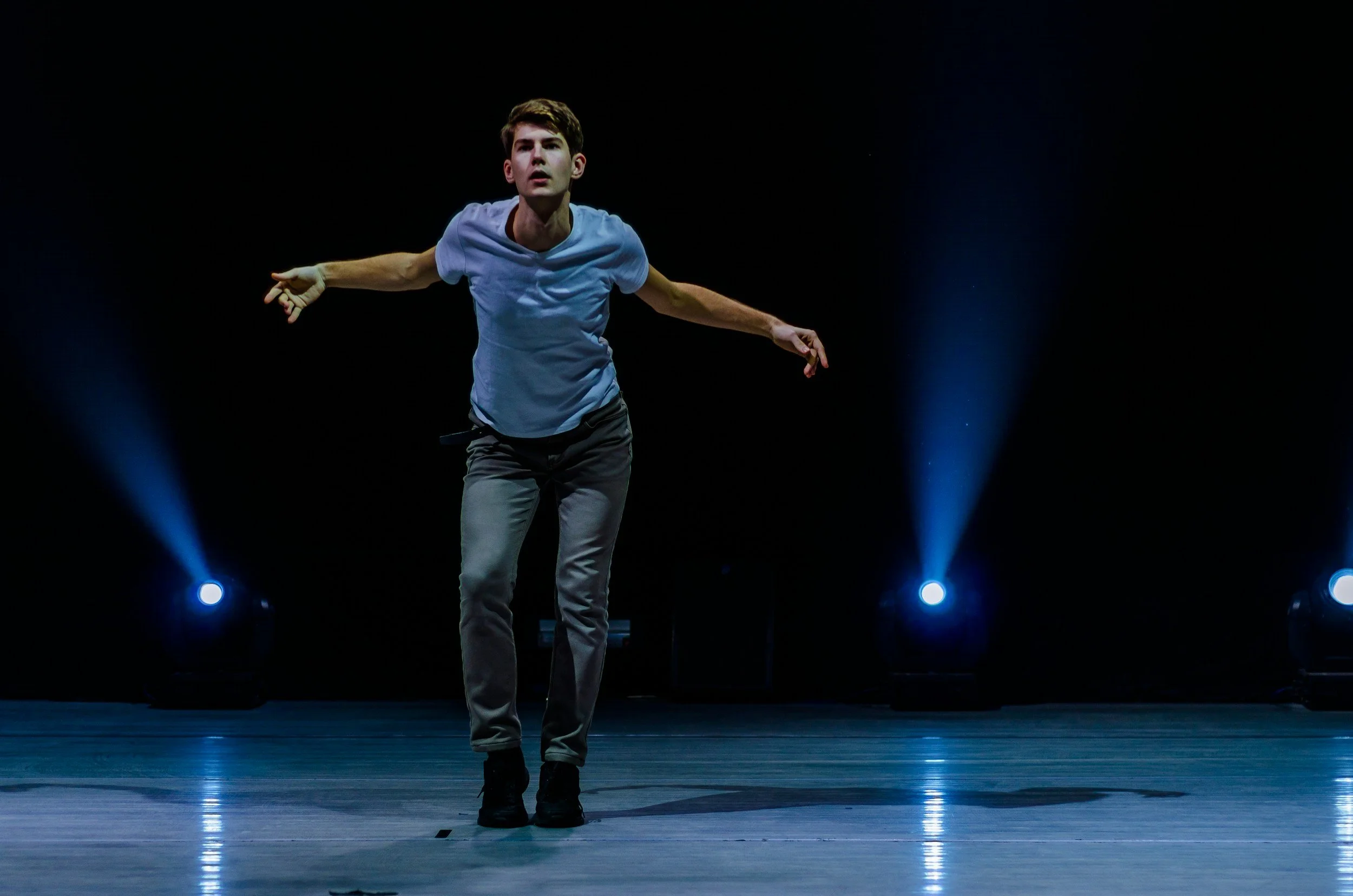 A young man performing a dance move on a stage with dark background and blue stage lights.