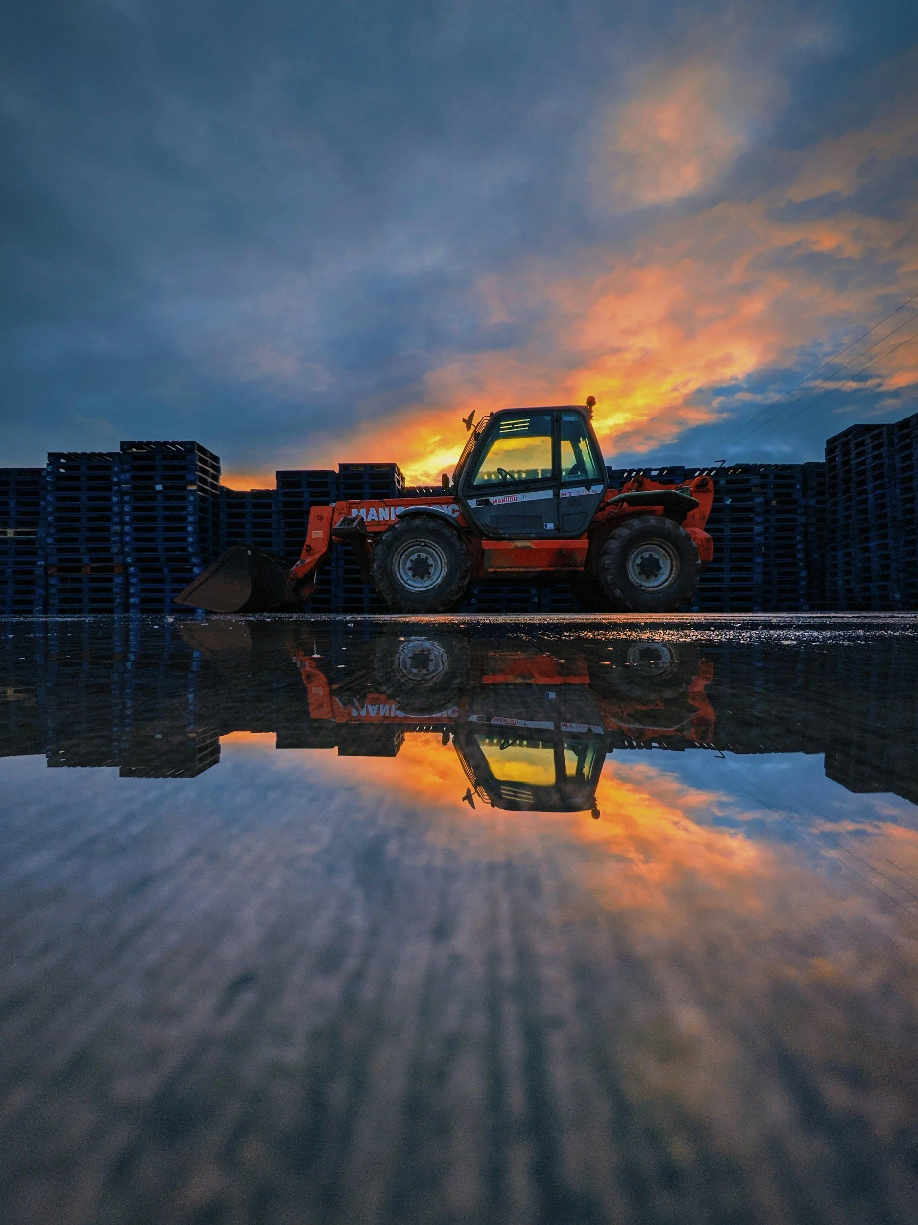 A construction loader at sunset with a reflected image on the wet ground.