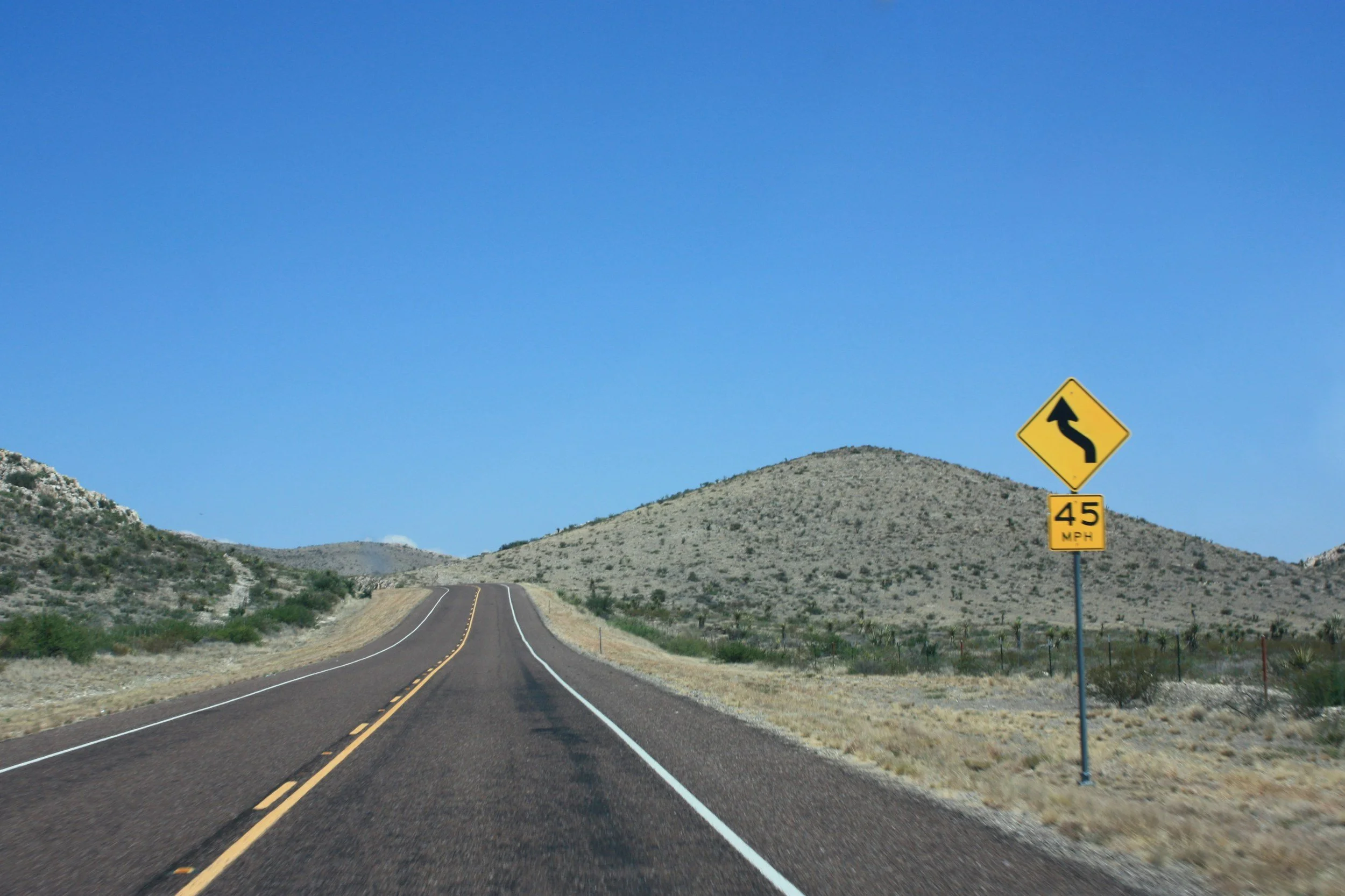 An empty two-lane road in a mountainous desert area with hills on either side and a clear blue sky.