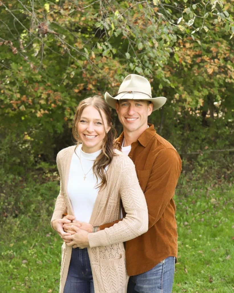 A smiling couple standing outdoors under green trees, with the woman in a beige cardigan and white turtleneck, and the man in a brown shirt and cowboy hat.