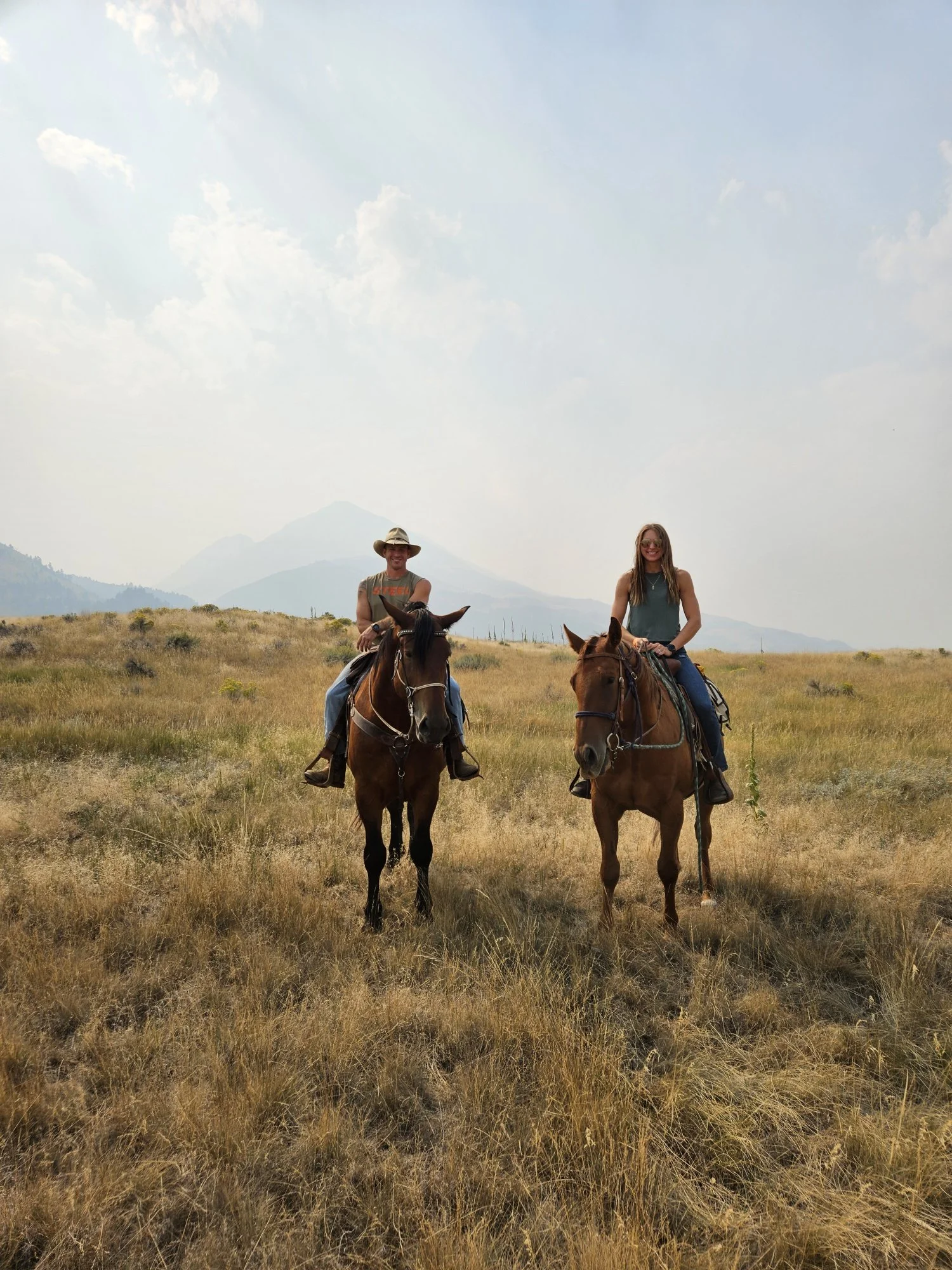 Two people riding horses in a grassy field with mountains in the background, under a partly cloudy sky.