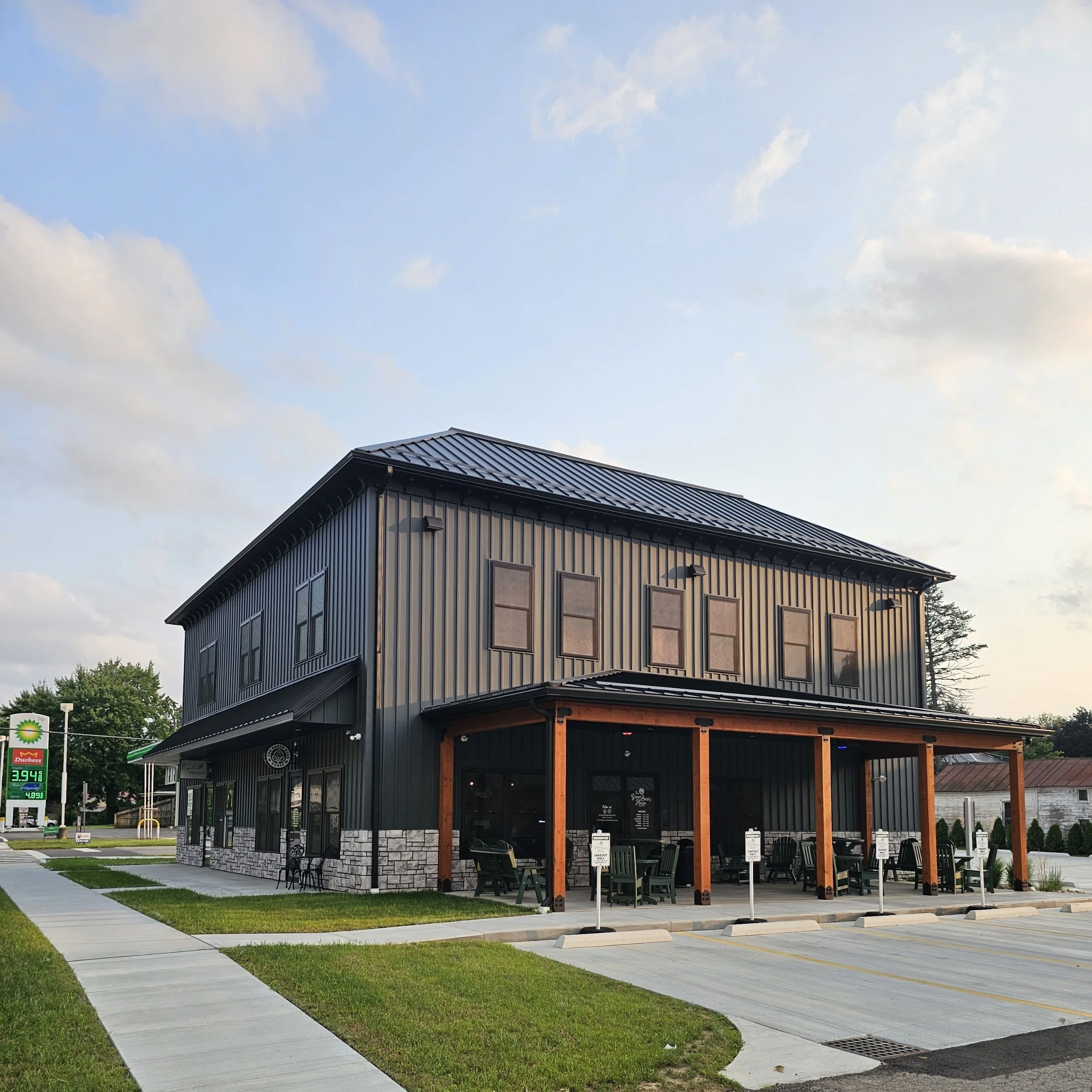 A modern two-story building with black metal siding and large windows, outdoor seating area with tables and chairs, a corner stone wall lower section, and a parking lot with a gas station sign showing fuel prices in the foreground.