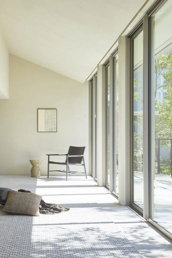 A sunlit modern living room with large floor-to-ceiling windows, a black chair, a small beige side table, a wall art piece, a pillow, and a blanket on a textured rug.
