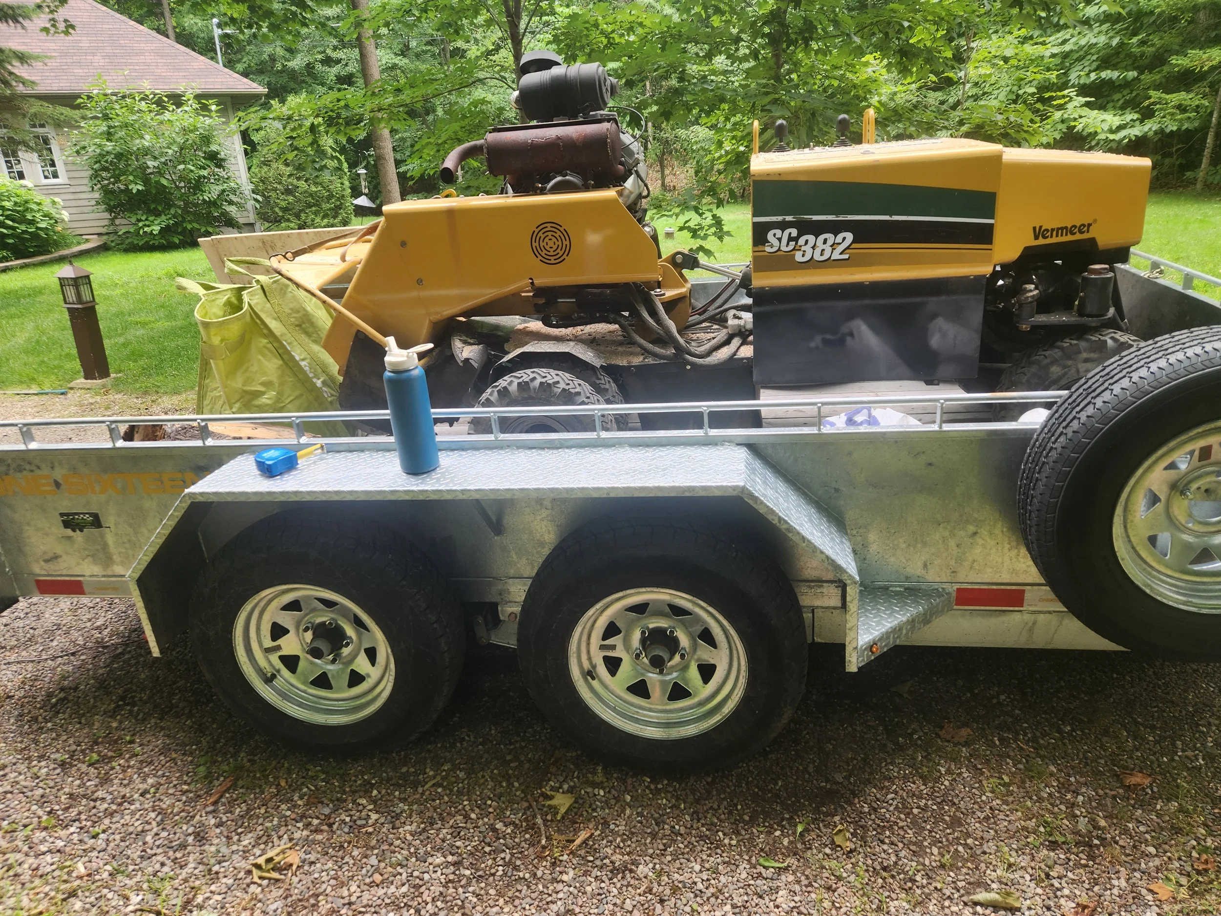 A yellow Vermeer SC382 wood chipper on a trailer, parked outdoors with a house and trees in the background.