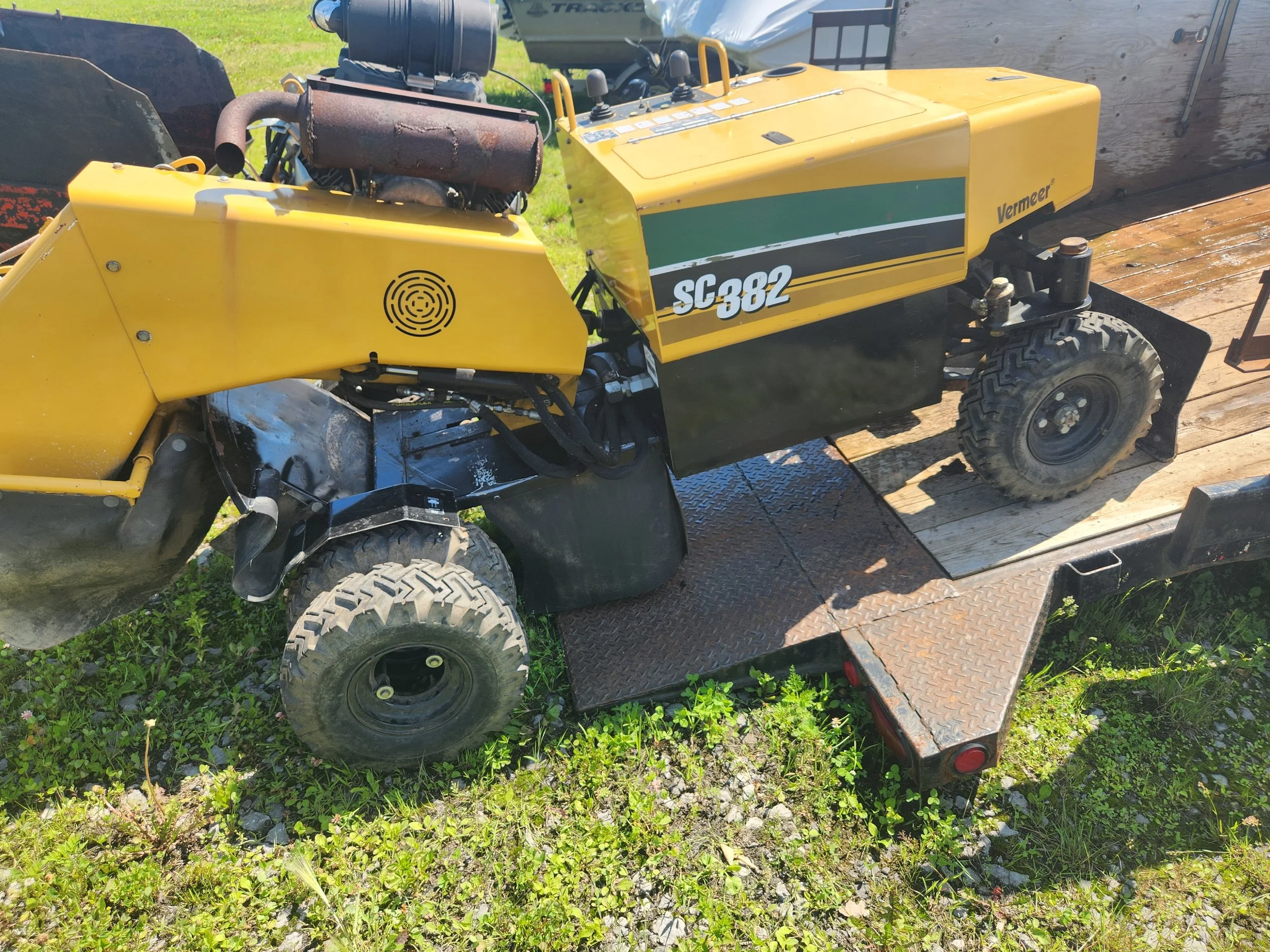 A yellow Vermeer SC382 stump grinder mounted on a trailer on grass and gravel.