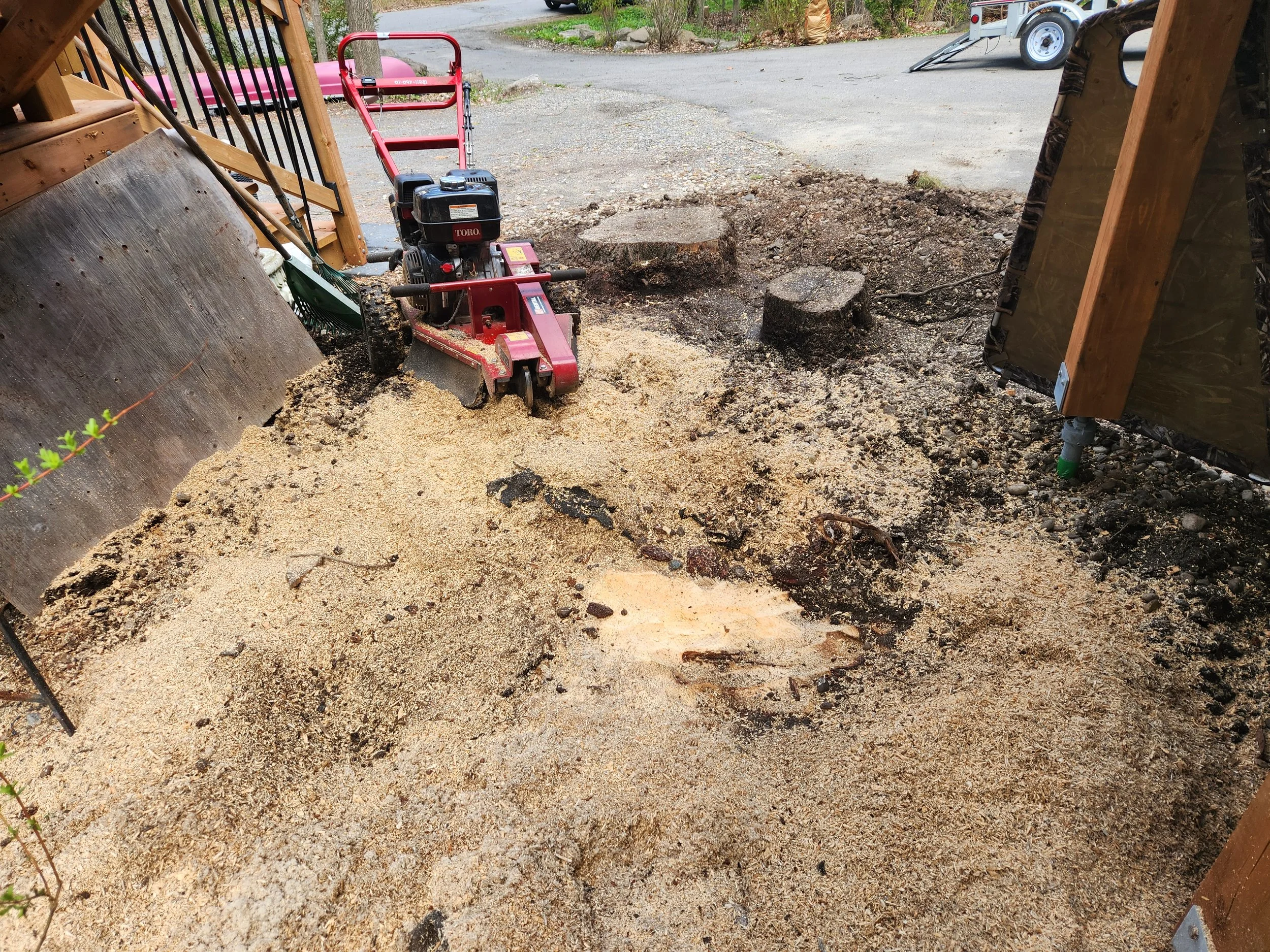 A small red and black mechanical plate compactor with a handle, used for compacting soil or gravel, positioned on a gravel surface outdoors surrounded by construction materials and equipment.