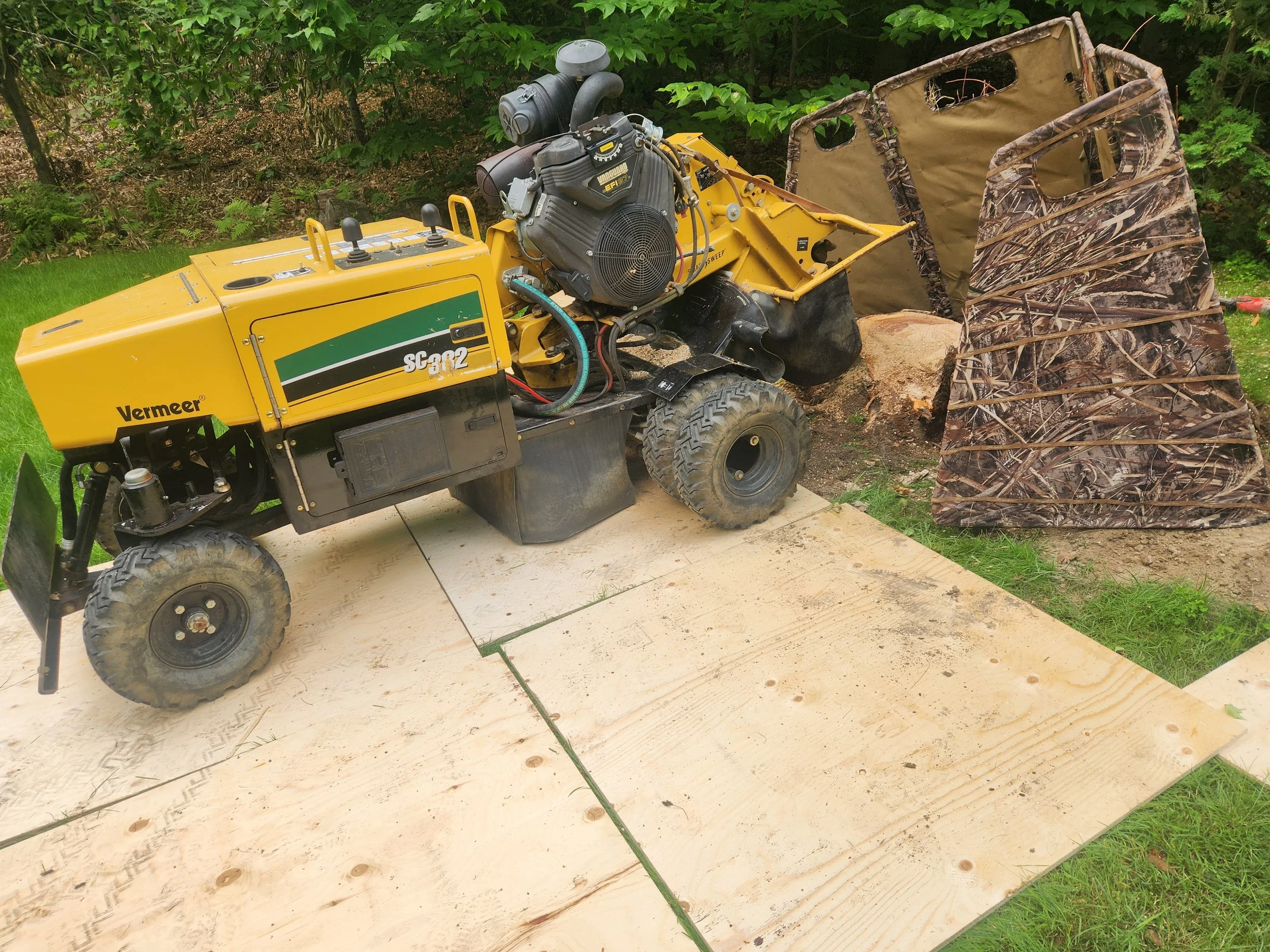 A yellow Vermeer stump grinder with black wheels and engine, positioned outdoors on a piece of plywood, with a pile of wood chips and a camouflage-patterned debris screen in the background.