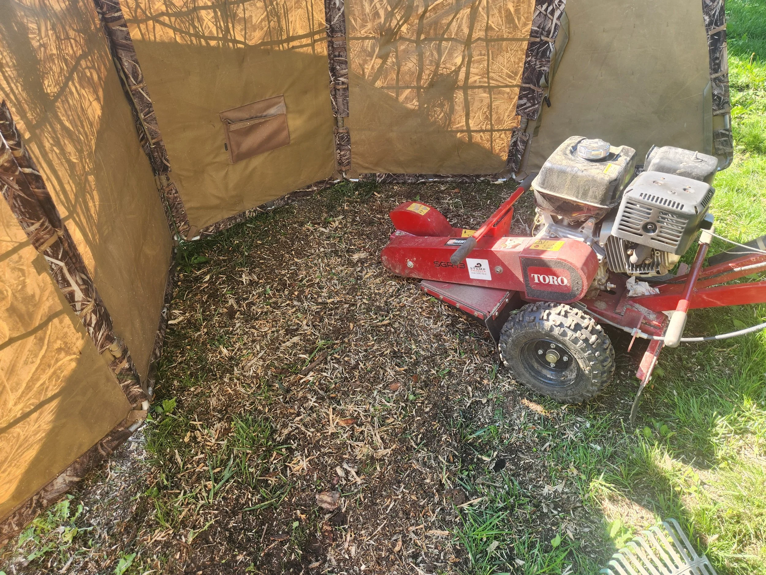 A red walking mower with an engine, small wheels, and a handlebar, positioned on grass and dirt, with a beige outdoor fabric structure nearby.