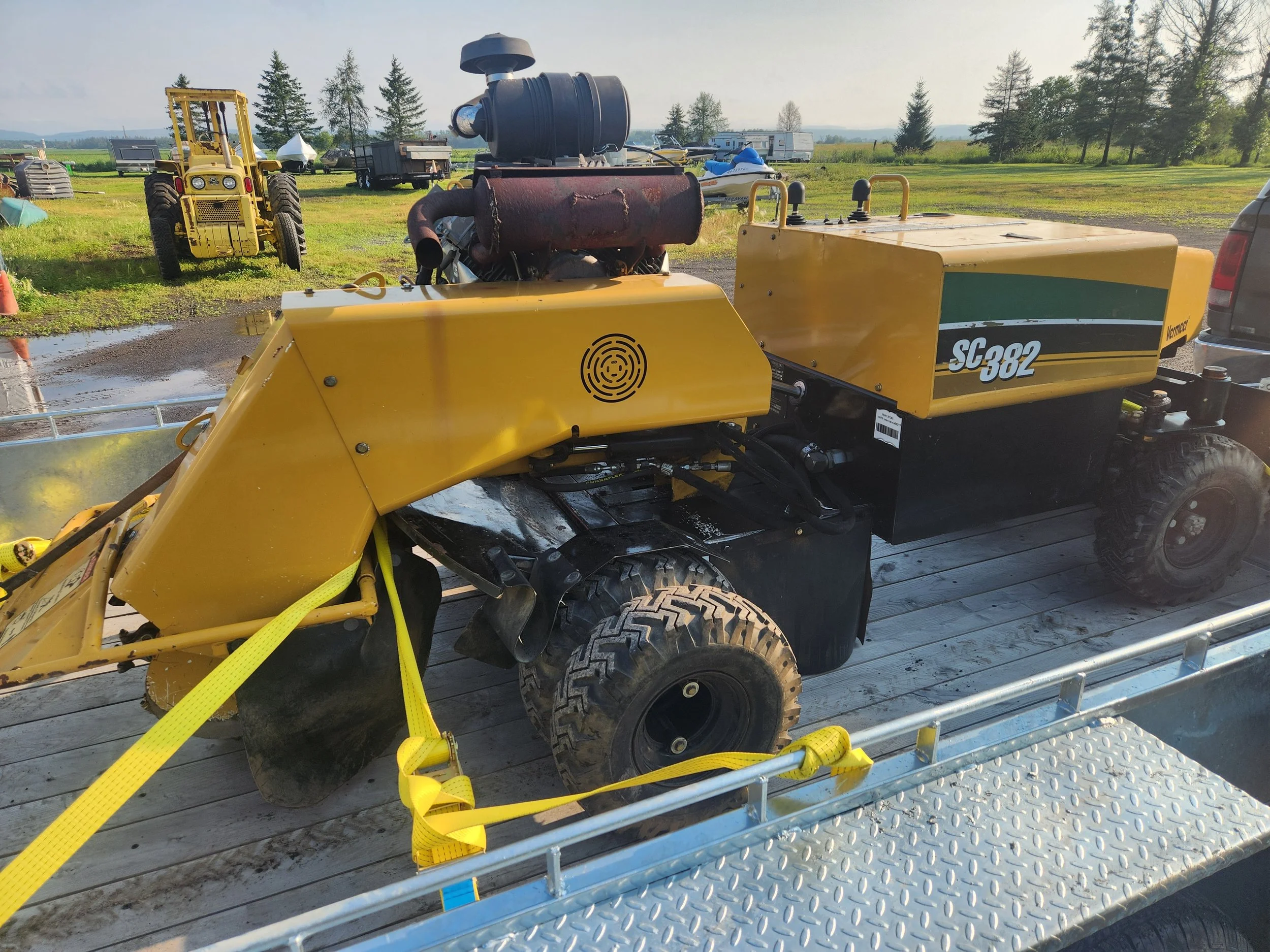 A yellow industrial machine with large black tires is loaded onto a metal flatbed trailer. The machine has a rusted red component on top and various ventilation and control features. There are yellow straps securing it to the trailer. In the backgrou