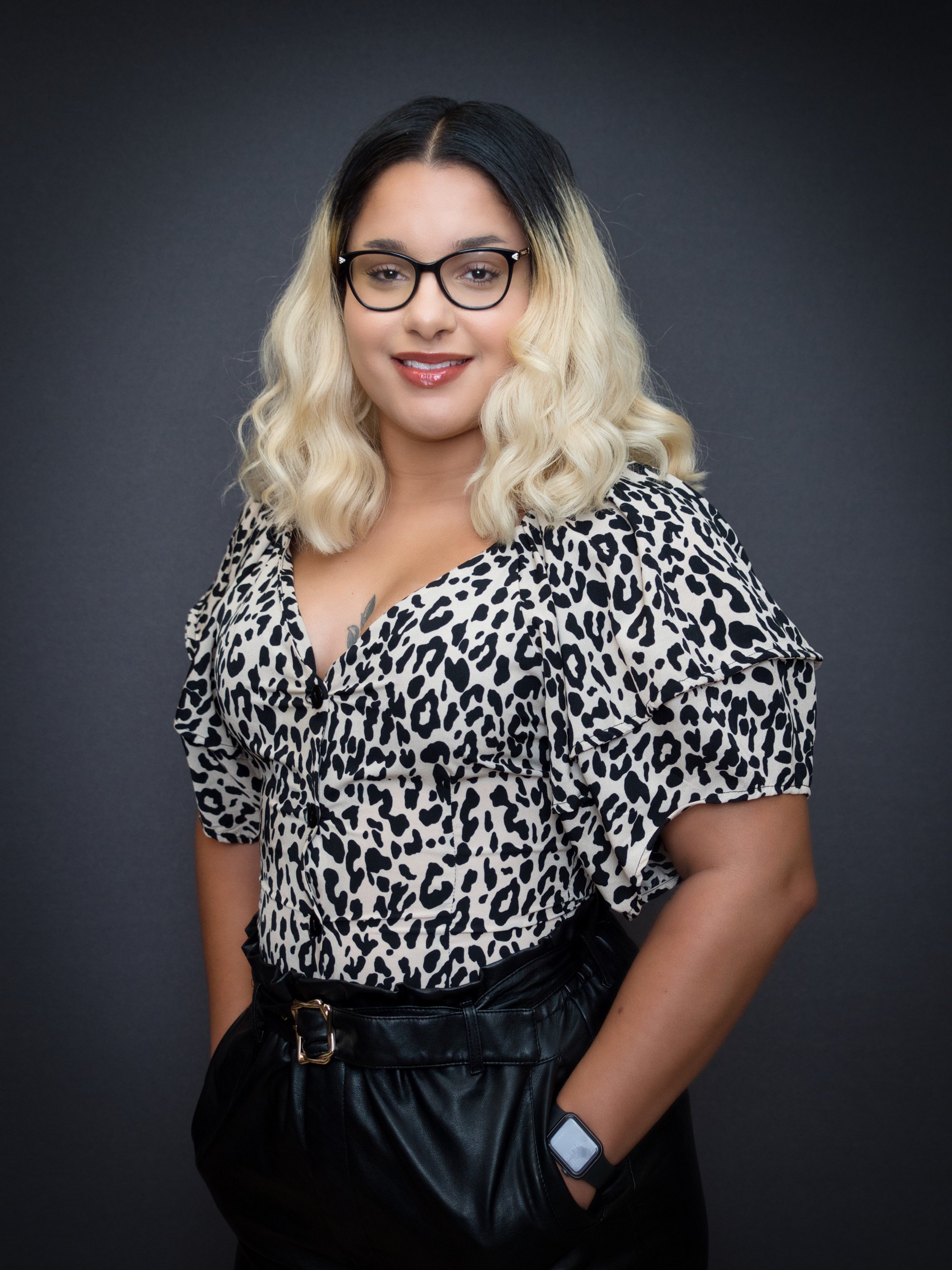 A woman with blonde hair, wearing black-rimmed glasses and a leopard print shirt, smiles at the camera against a dark gray background.