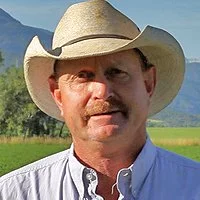 Marty Nieslanik wearing a wide-brimmed cowboy hat standing outdoors with a green field and mountains in the background.