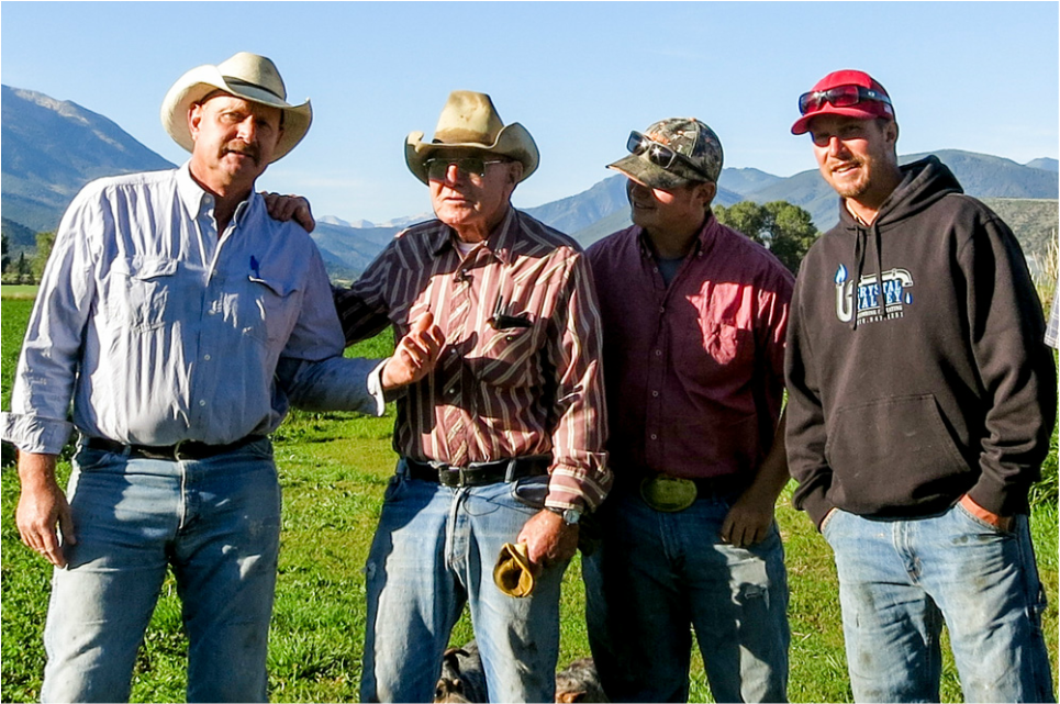 Four men standing outdoors in a grassy area with mountains in the background, dressed in casual and cowboy attire, including hats, sunglasses, and a hoodie.
