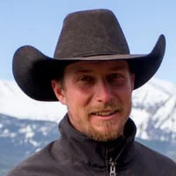 A man outdoors wearing a black cowboy hat and a black jacket with a mountain background.