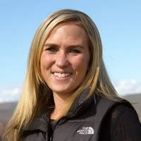 A woman with long blonde hair, smiling, wearing a black North Face jacket, outdoors with a cloudy sky