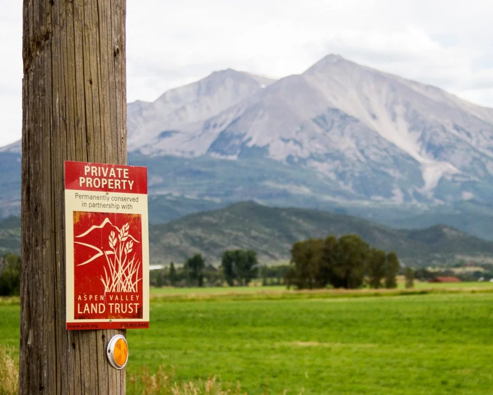 A wooden utility pole with a red and white sign that reads 'Private Property, Aspen Valley Land Trust, Permanently conserved in partnership with.' In the background, mountains and green fields are visible under a cloudy sky.