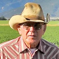 John Nieslanik wearing glasses and a cowboy hat, standing outdoors in a green field with mountains in the background.