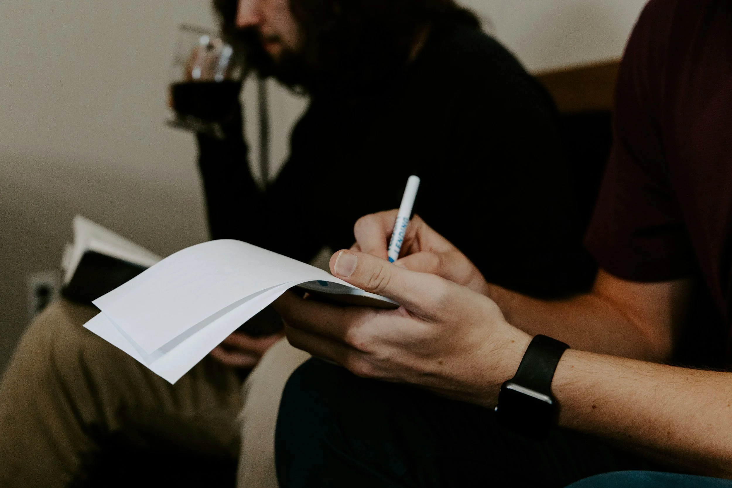 Person holding papers and a pen, with someone sipping from a glass in the background.
