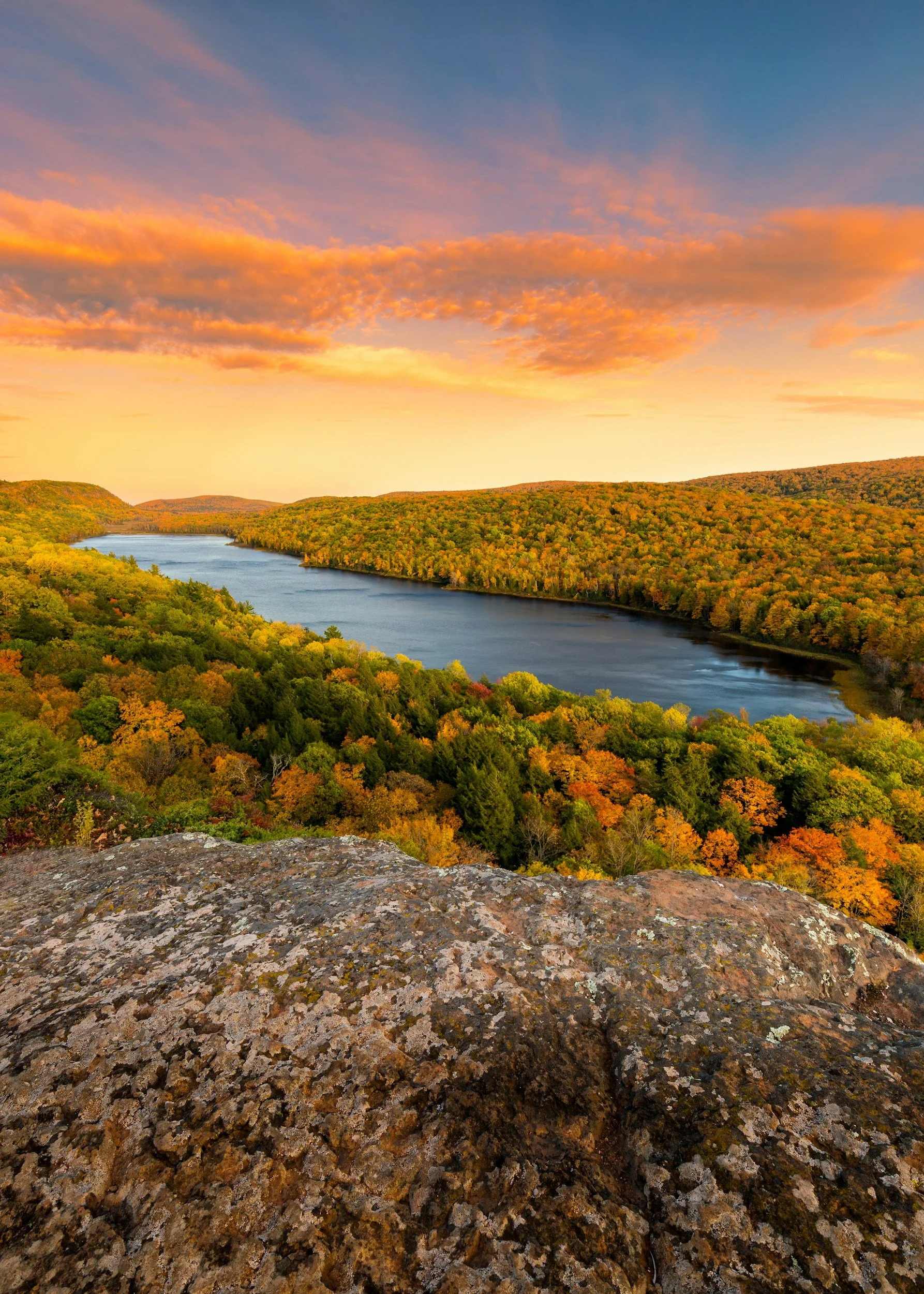 View from a rocky ledge overlooking a river winding through an autumn forest with colorful trees and a sky with pink and orange clouds at sunset.