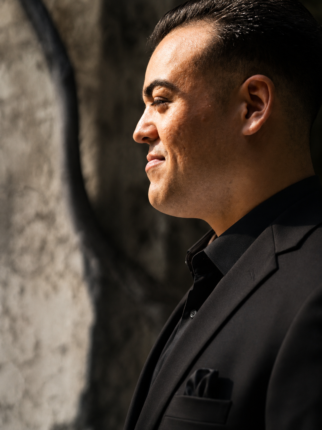 Side profile of a man in a black suit smiling in front of a textured wall.
