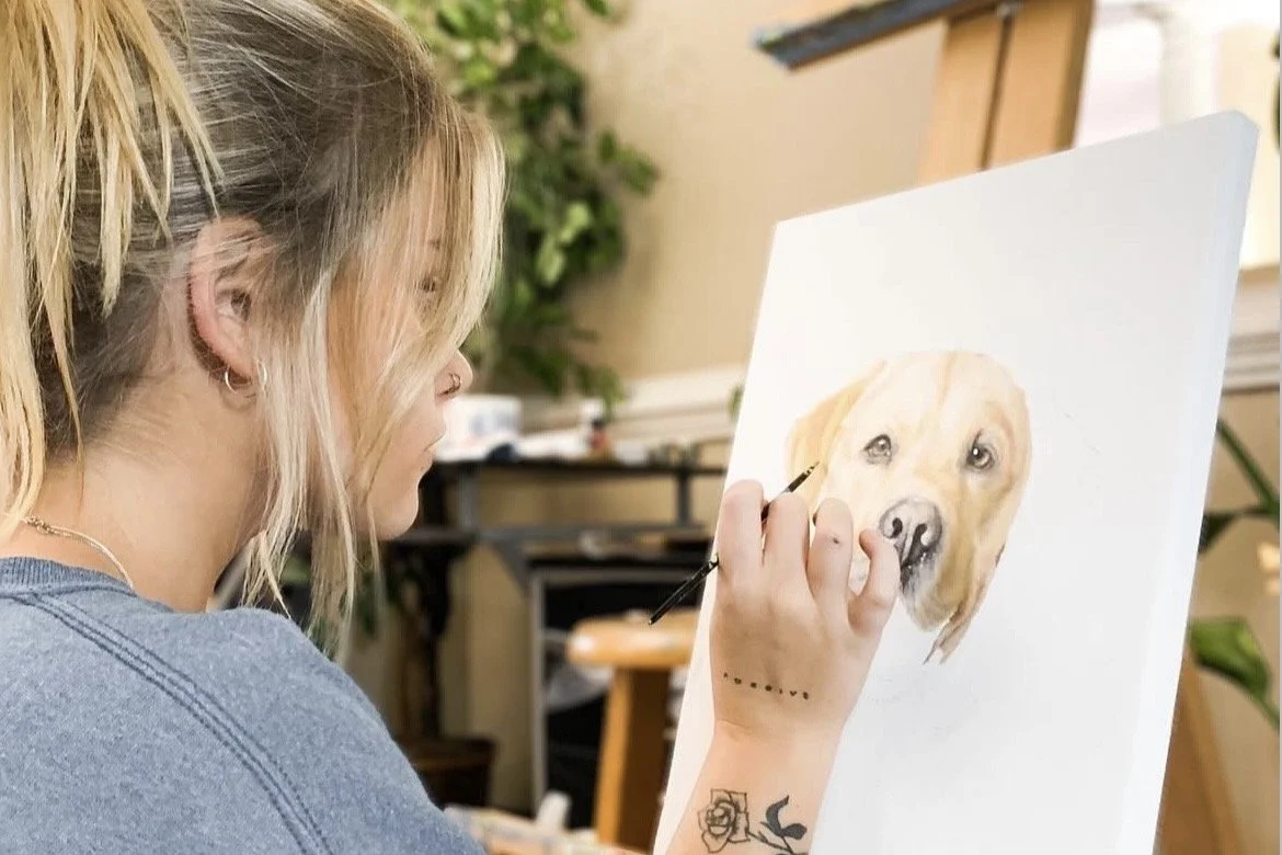 A woman with blonde hair and tattoos on her arm is painting a realistic portrait of a yellow Labrador retriever dog on a canvas in an art studio.