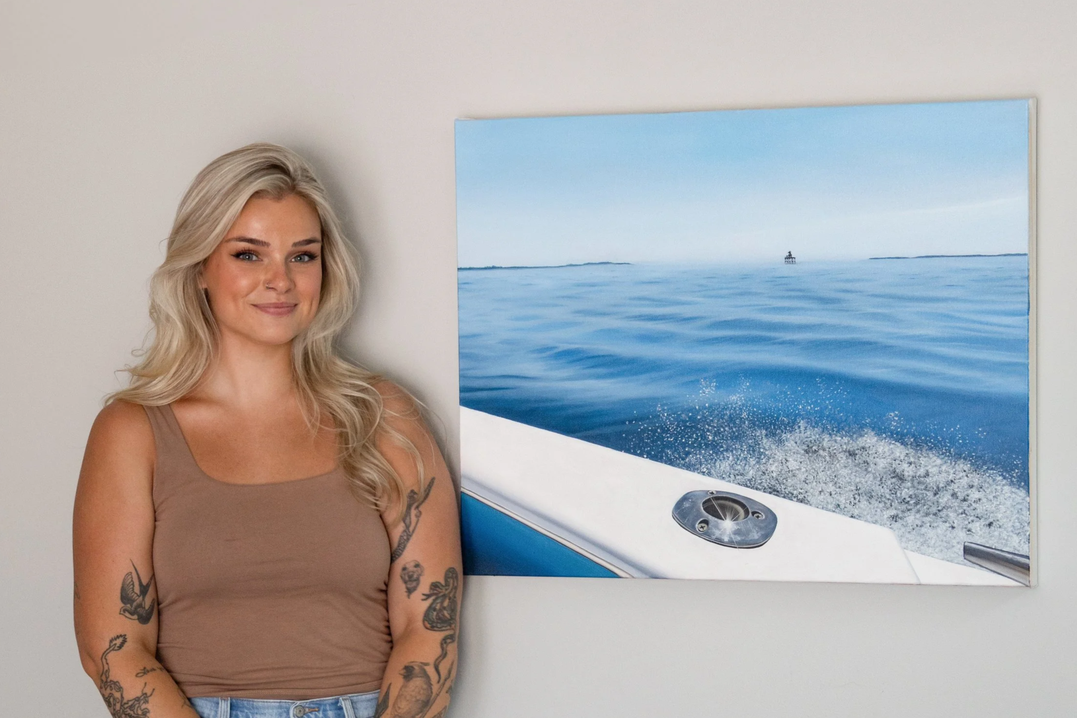 A young woman with blonde hair and tattoos on her arms standing next to a picture of a seascape with a boat in the distance, on a plain wall.