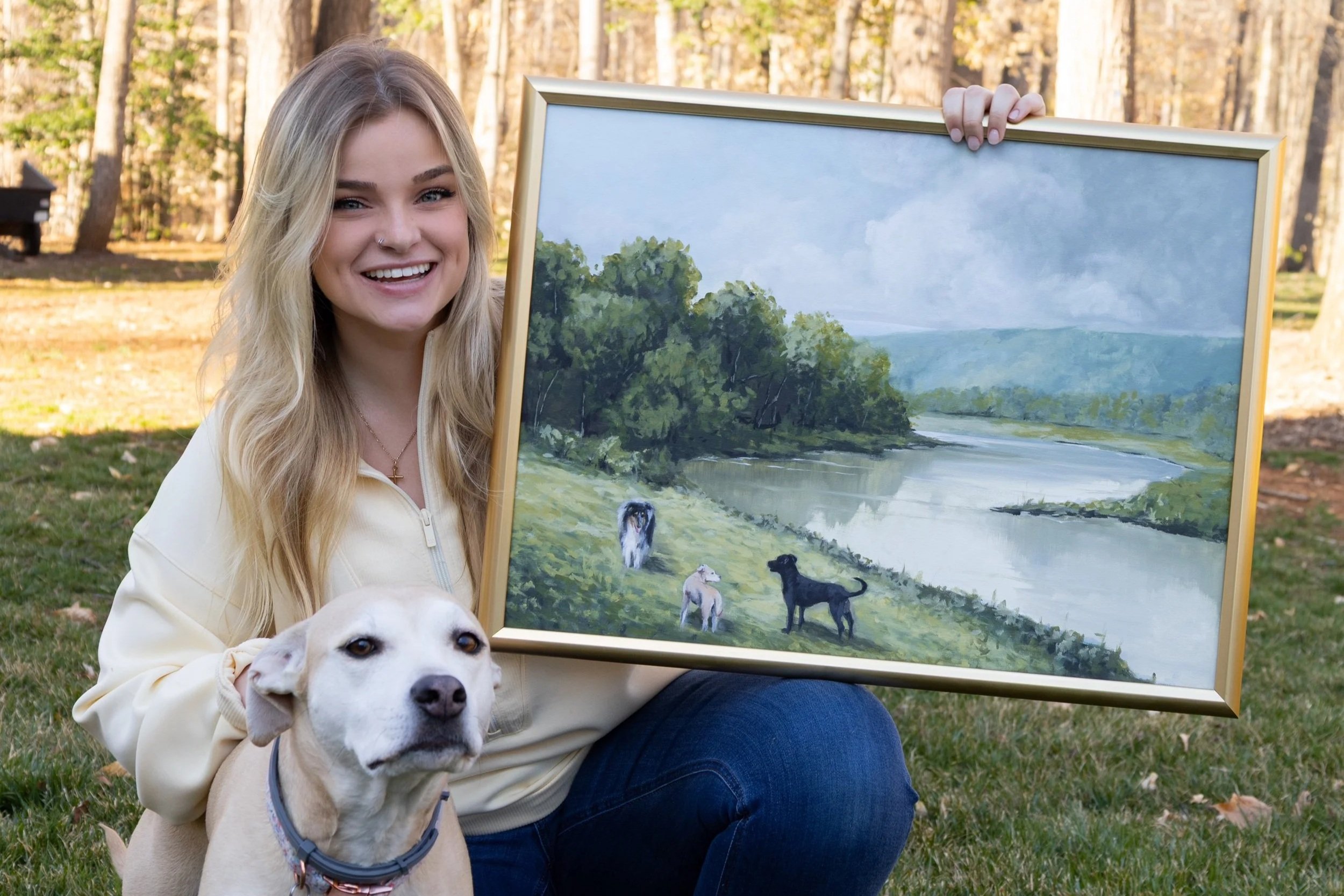 A smiling young woman with long blonde hair, wearing a beige jacket, sitting outdoors in a park, holding a framed landscape painting of a river, trees, and a cloudy sky. A white dog with a collar is sitting in front of her, partially blocking her lap