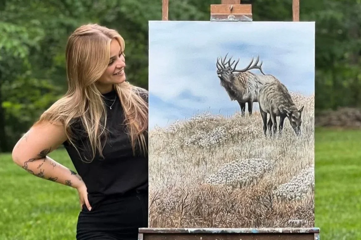 Artist standing outdoors next to her completed painting of a majestic elk in a field.