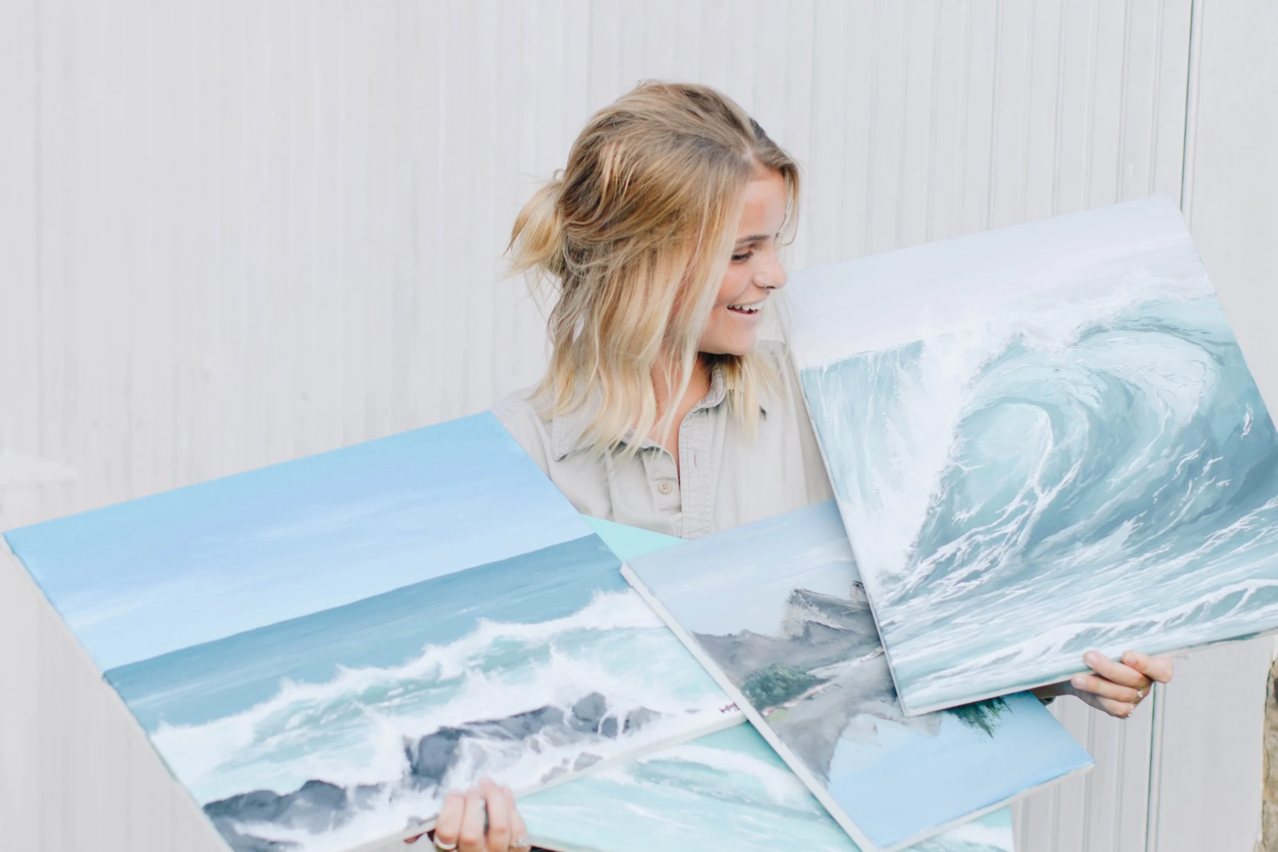 A person with blonde hair smiling while holding up water-themed paintings of waves and ocean scenes.