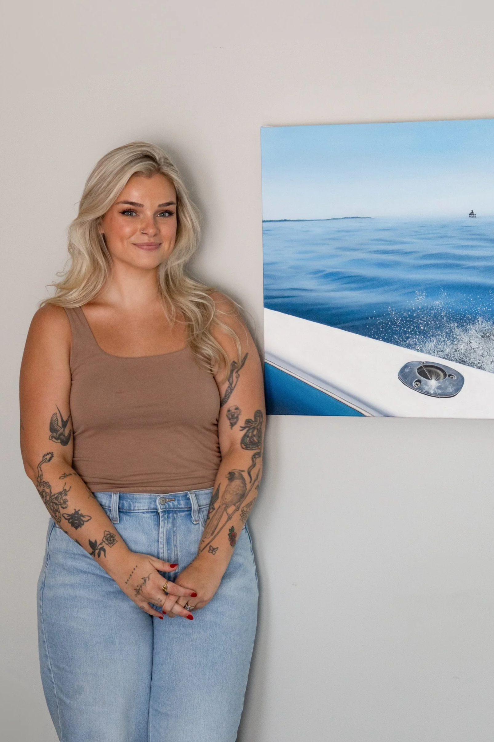 A young woman with blonde hair and tattoos on her arms is standing against a white wall next to a framed picture of a boat on the water.