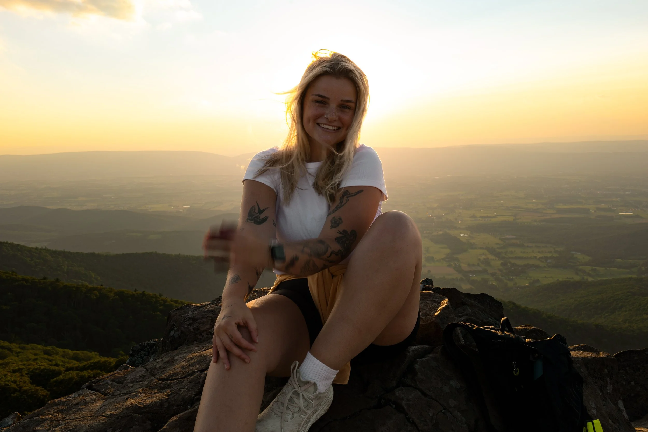 A young woman with blonde hair, tattoos, and a white t-shirt sitting on a mountain summit during sunset, smiling at the camera with a panoramic view of the landscape below.