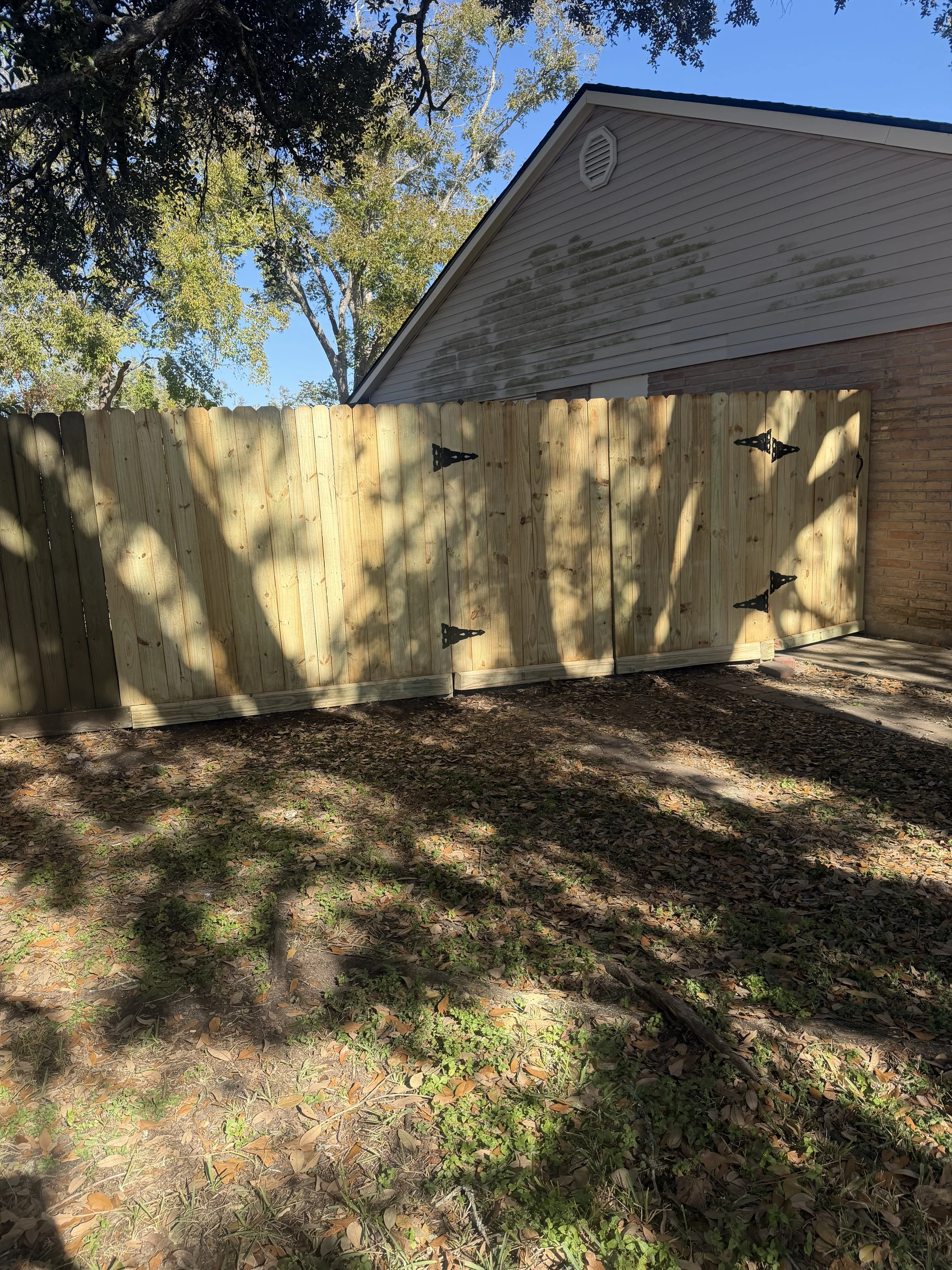 Wooden fence with decorative black hinges in a backyard, trees with green leaves, and a house with a gray roof and vents in the background.