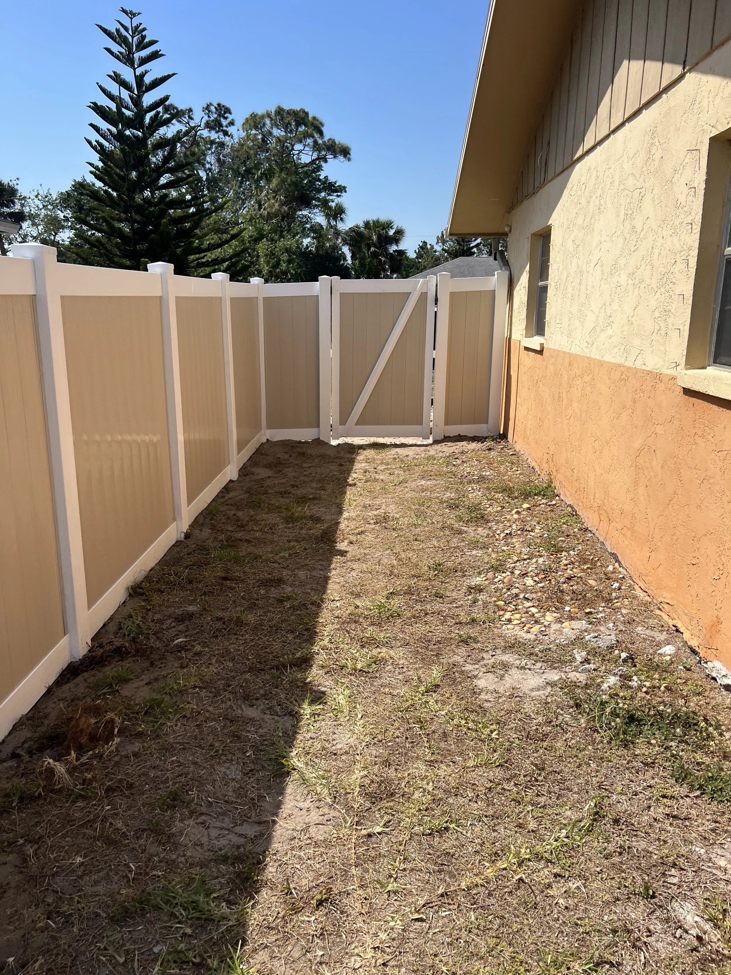 Side yard with dirt and sparse grass, beige vinyl fence, yellow stucco house with orange lower wall, two windows, and an overhanging roof, tall trees in the background, clear blue sky.