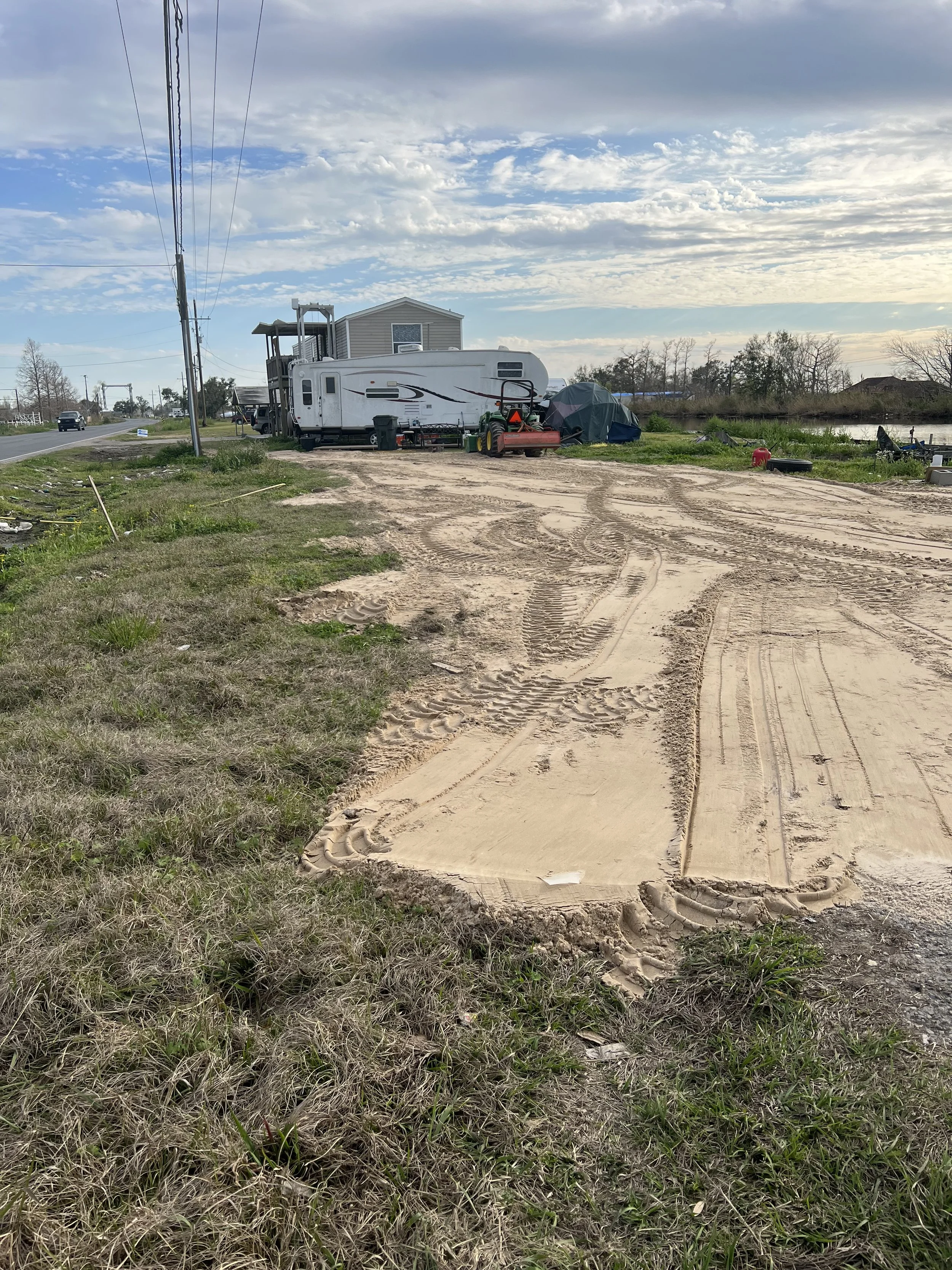 A sandy area near a body of water with a trailer, equipment, and a tent. In the background, there are trees, a river, and a cloudy sky.