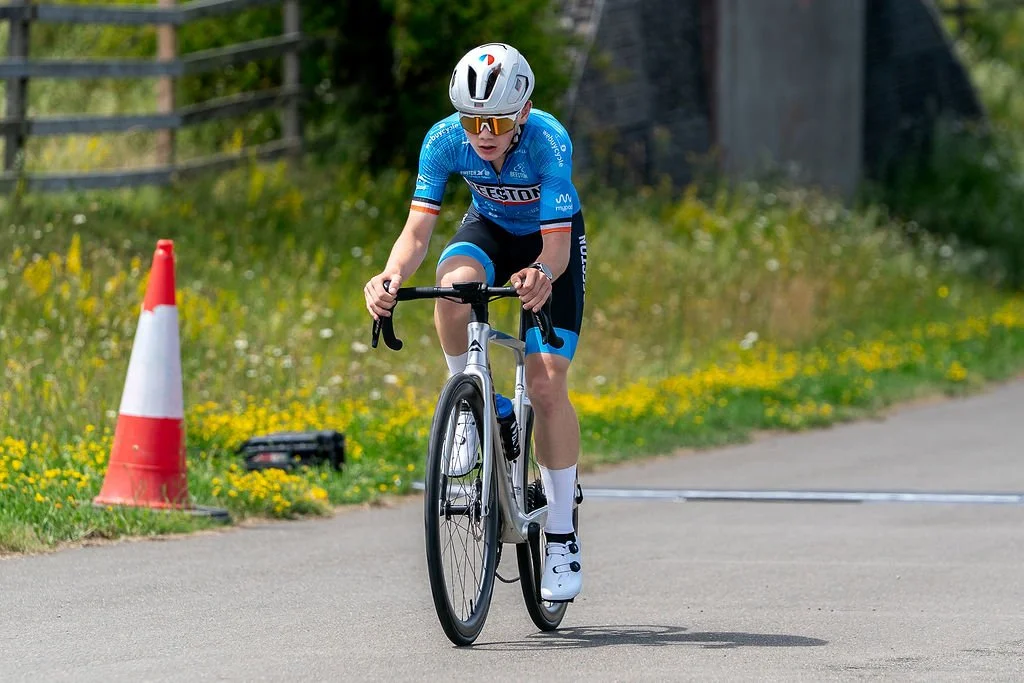 A young male cyclist wearing a blue racing jersey, black shorts, white helmet, and sunglasses riding a bike on a paved road next to a traffic cone, with a grassy area and a wooden fence in the background.