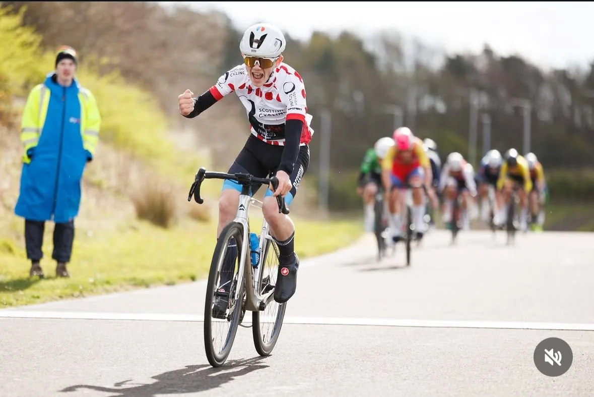 A cyclist celebrating as they lead a bike race on a countryside road, with a group of racers in the background and a person watching on the side.