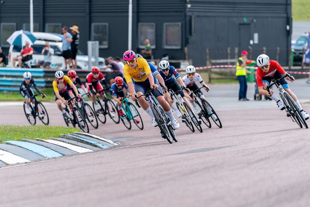 A group of young cyclists racing on a track, leaning into a turn, with spectators watching in the background.