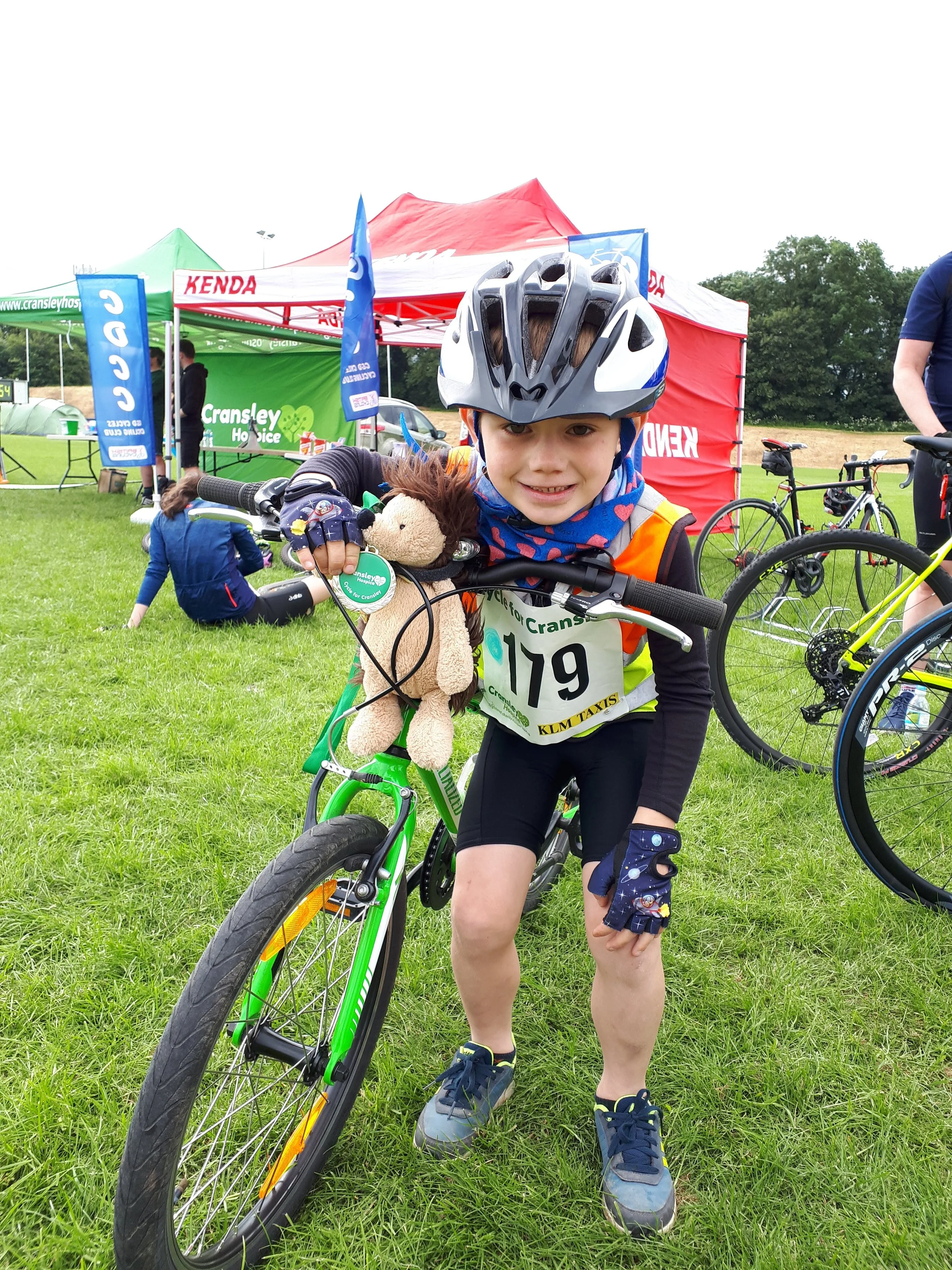 A young boy wearing a safety helmet and gloves, smiling, standing next to a green bicycle during a cycling event. He has a stuffed animal toy hanging from his bike handlebar. Behind him are tents and banners with cycling-related logos, and other participants and bicycles are visible on the grassy field.