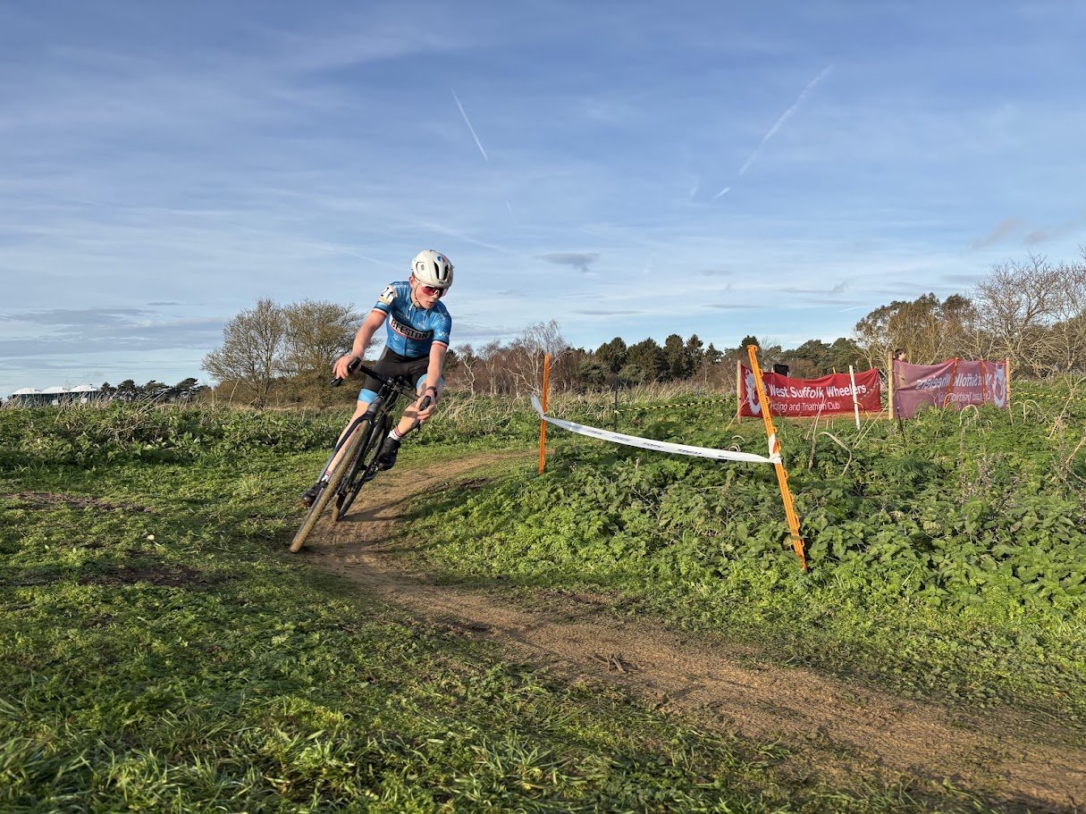 A person riding a mountain bike on a dirt trail during daytime, wearing a helmet and sports gear, with trees and banners in the background.