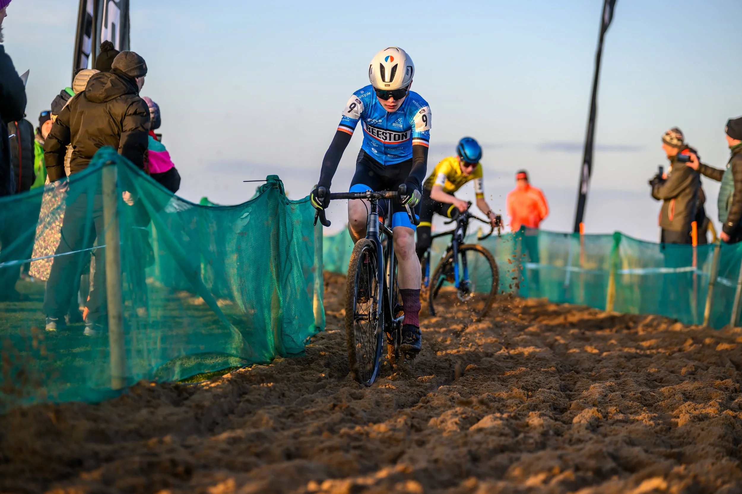 Cyclist in blue jersey and helmet riding mountain bike through a dirt course with spectators watching.