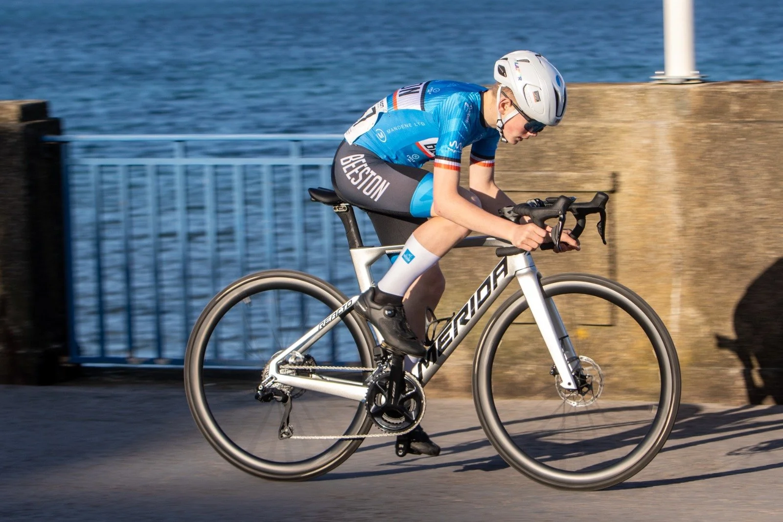 A cyclist wearing a blue and black racing outfit and white helmet riding a white Merida road bike along a waterfront path.