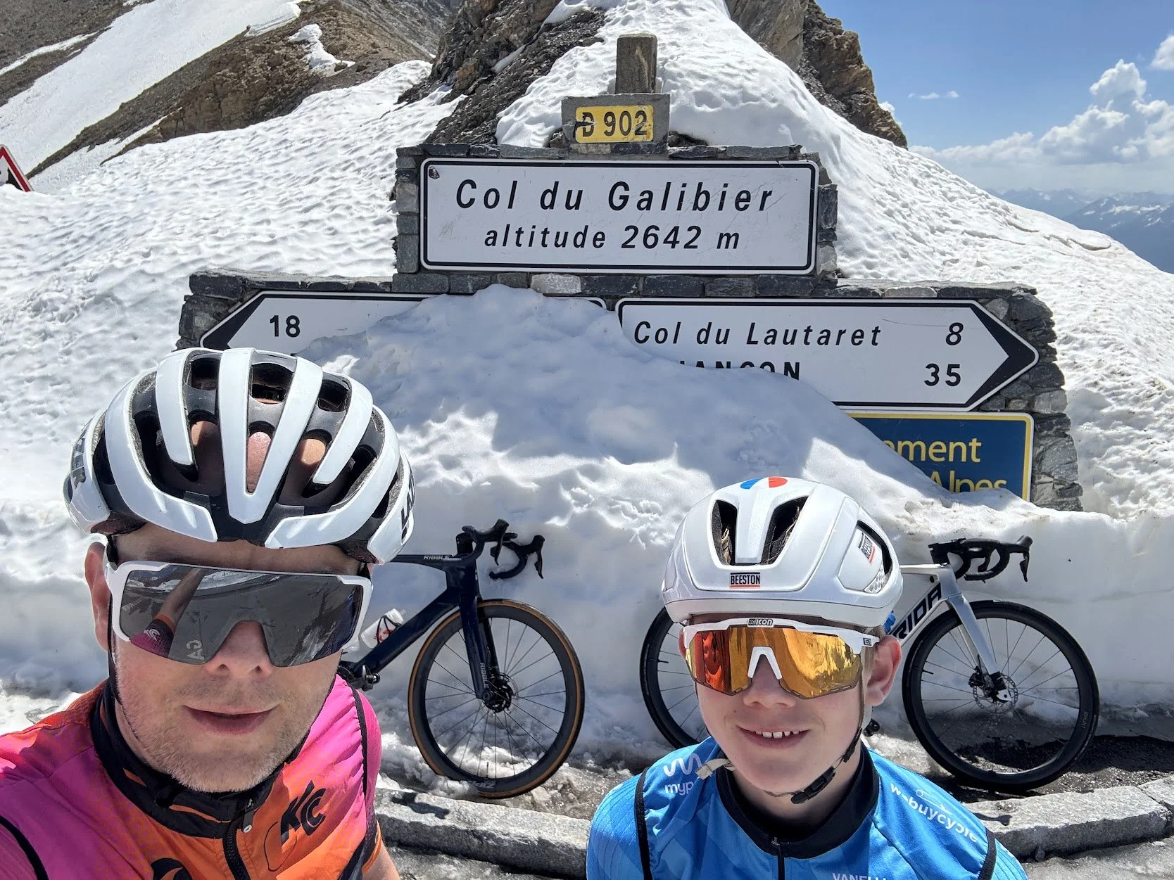 Two cyclists taking a selfie in snowy mountain terrain with snow-covered peaks in the background, posing near a sign indicating the Col du Galibier at an altitude of 2642 meters, along with bicycles